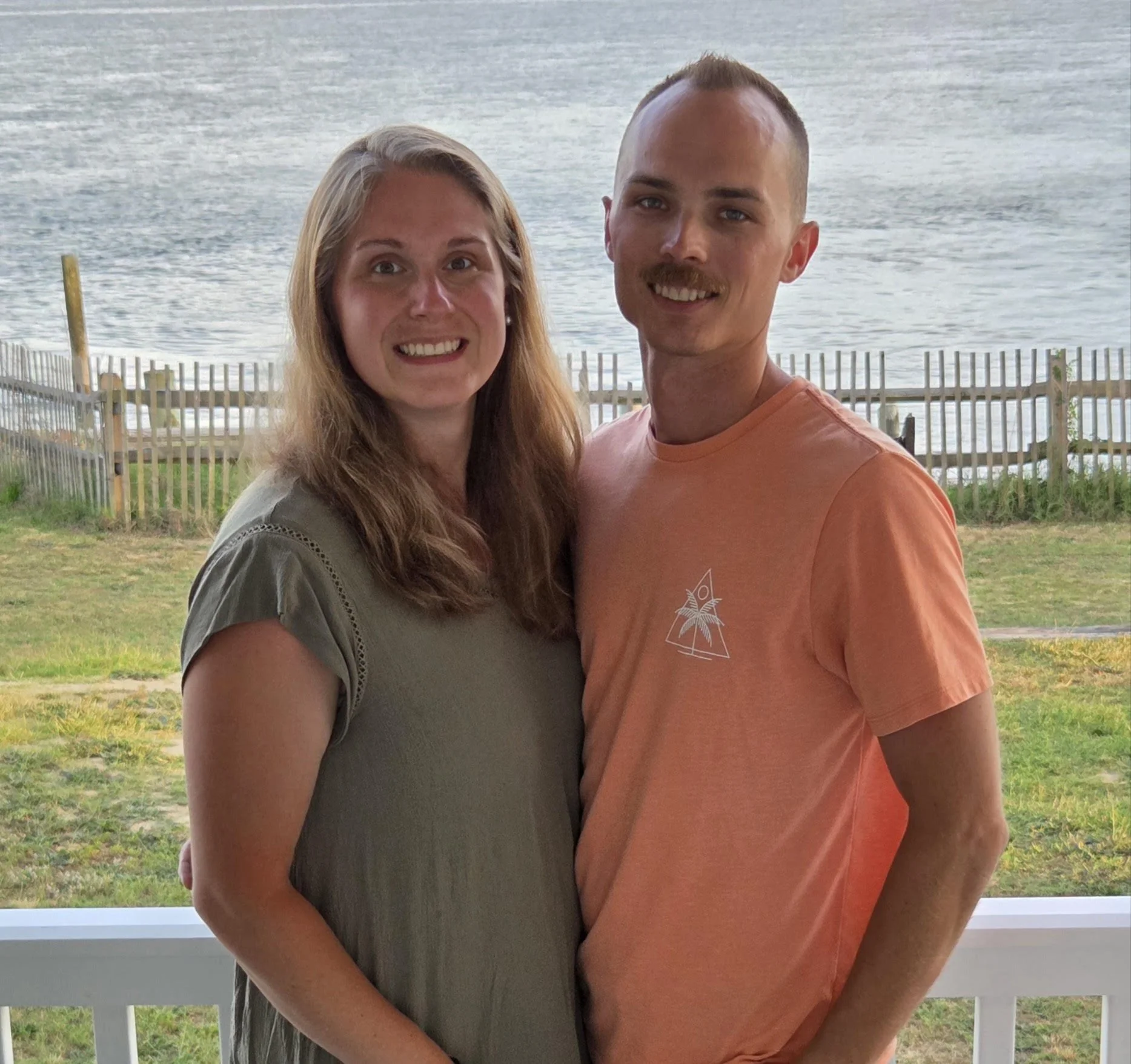 A smiling woman with long blonde hair and a man with short hair and a mustache, standing outdoors near water with a wooden fence in the background.