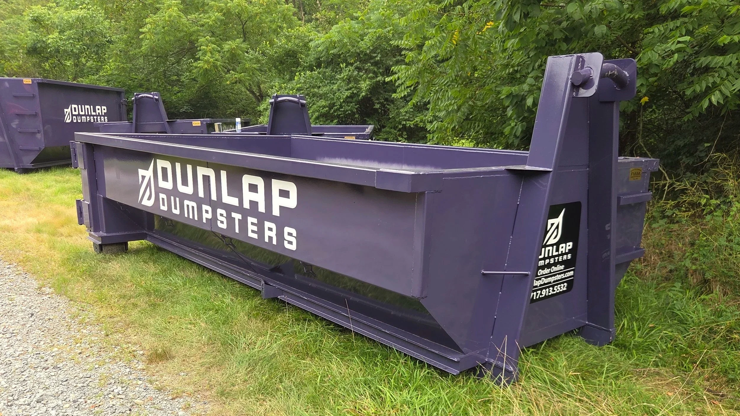 Large purple dumpster with white logo and text for Dunlap Dumpsters, set on grass and gravel near trees.