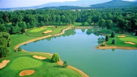 A golf course with green fairways, sand bunkers, and a large water hazard, surrounded by trees and distant mountains.