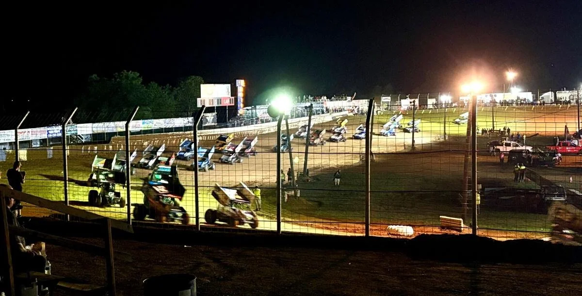 Nighttime dirt track racing event with multiple race cars on the track, bright floodlights, and spectators behind a safety fence.