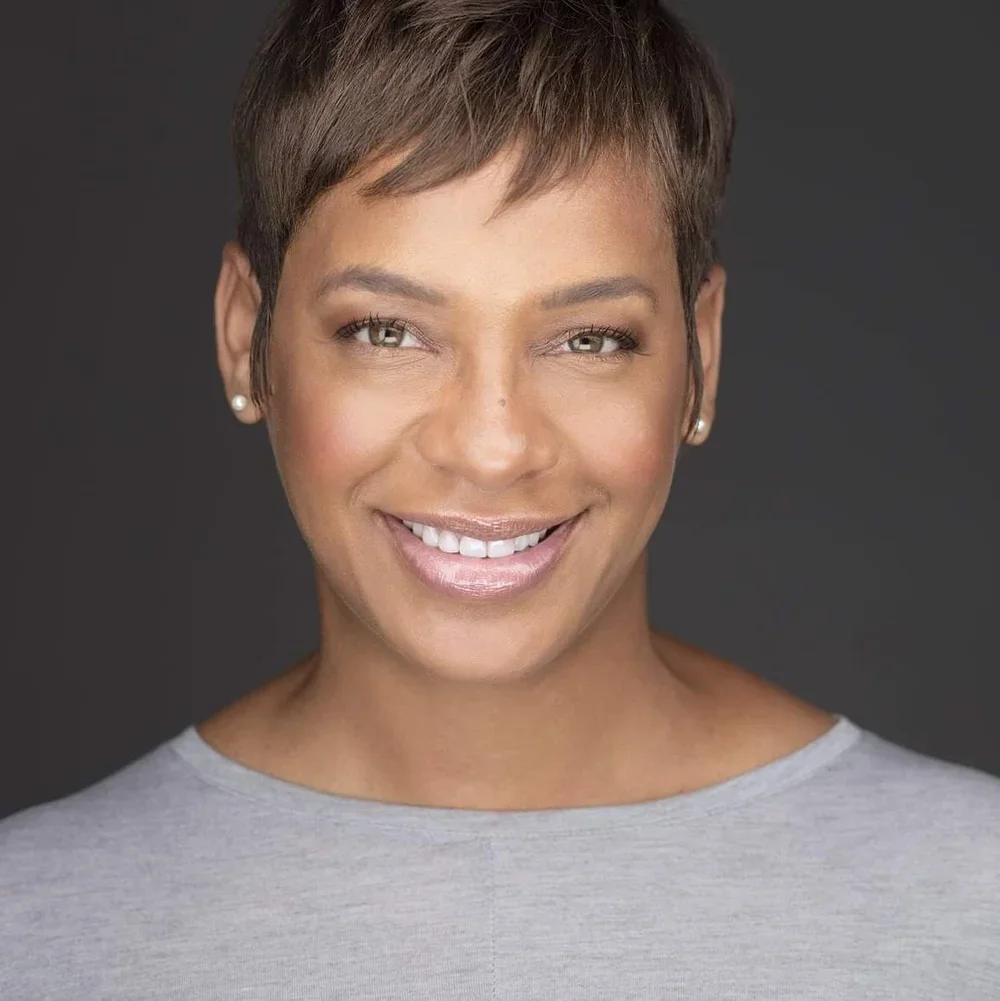 A woman with short brown hair styled in a pixie cut, smiling, wearing a light gray top and pearl earrings, against a dark background.