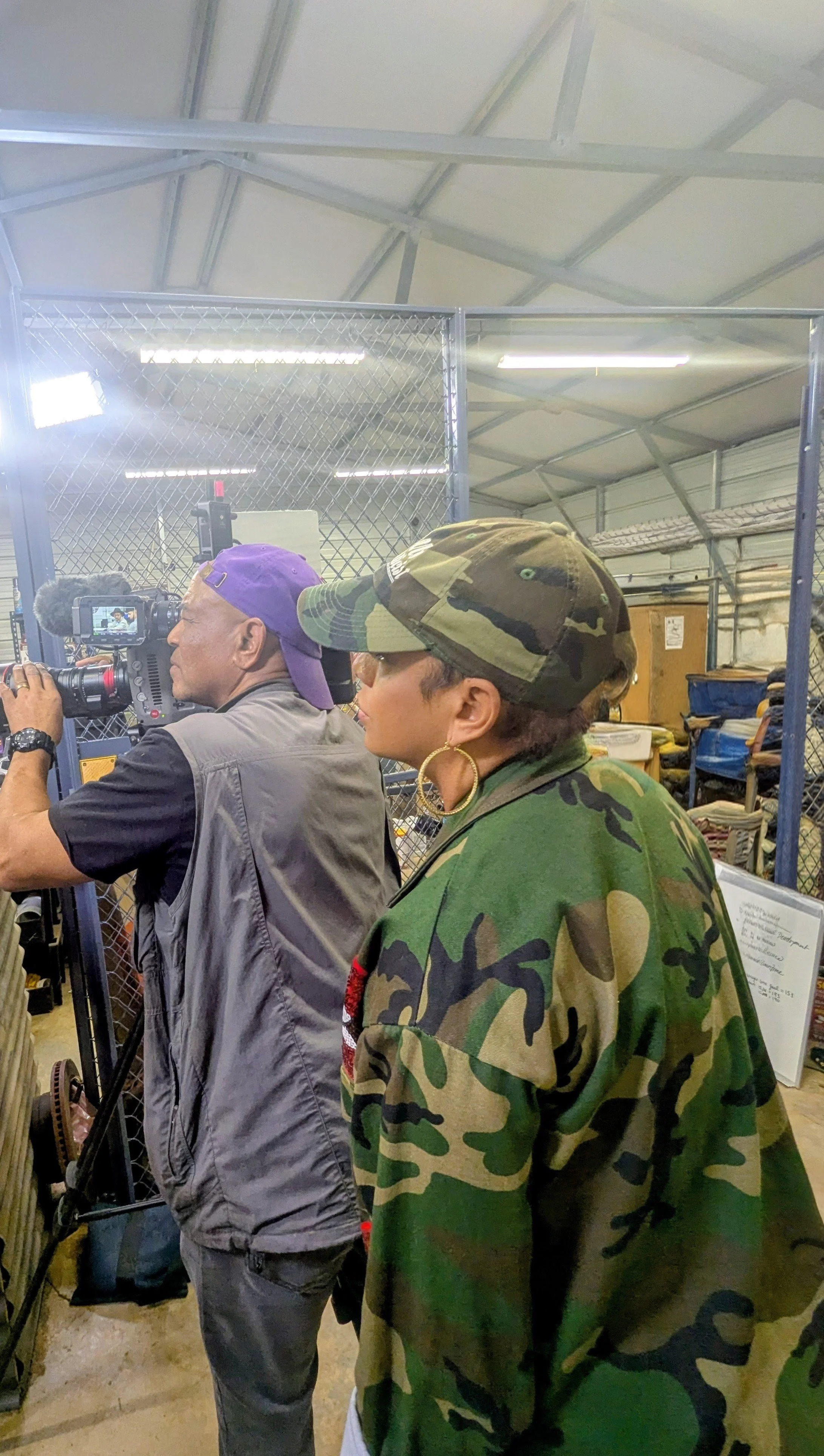 Areshia McFarlin with short dark hair, wearing large gold hoop earrings, glasses, a camouflage jacket, and a matching camouflage cap, is observing a man filming with a video camera at an indoor location with metal fencing and equipment.