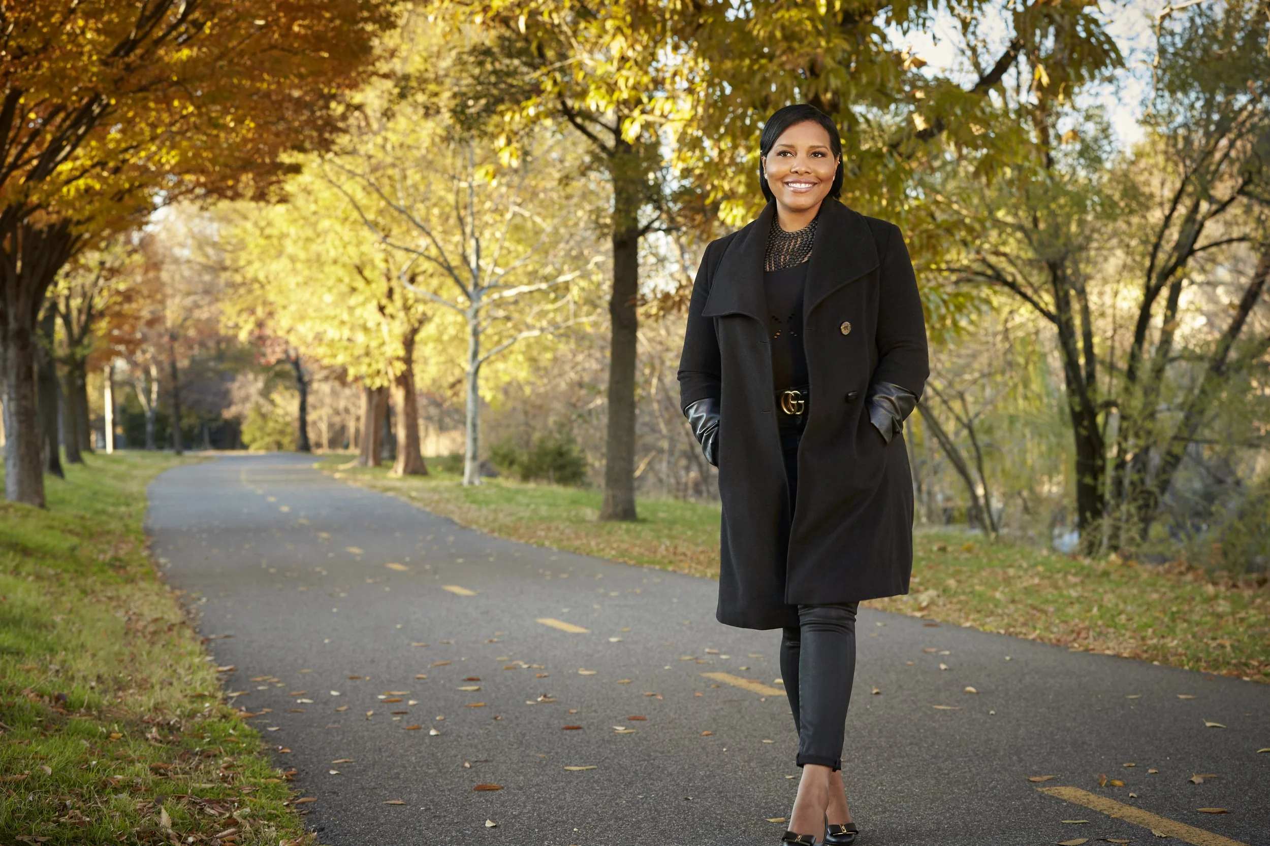 A woman walking on a curved paved path in a park with autumn-colored trees, wearing a black coat, black pants, and high heels, smiling.