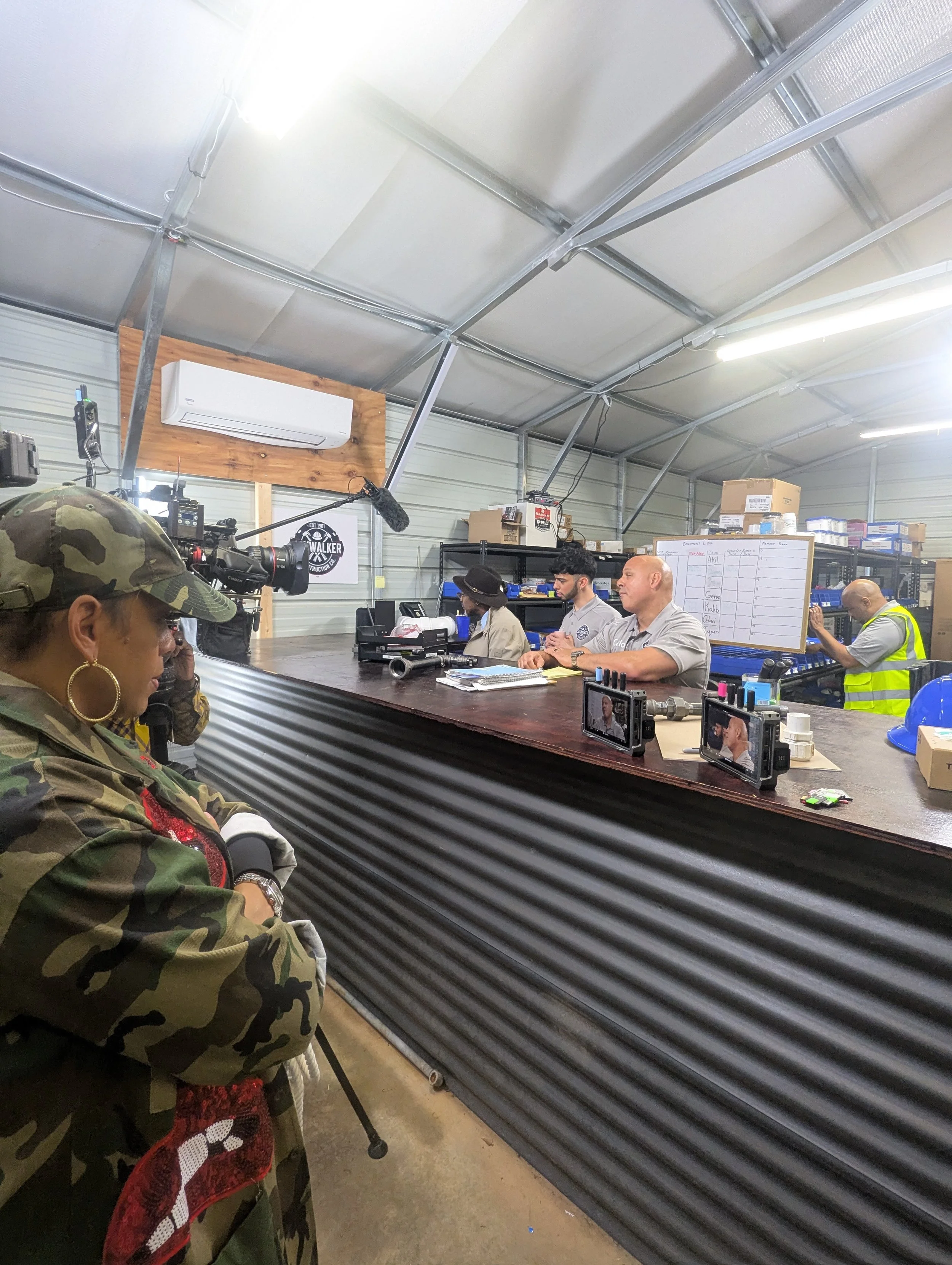 People seated at long desk in a warehouse, with a camera crew filming them. The warehouse has an air conditioning unit and a whiteboard with notes.