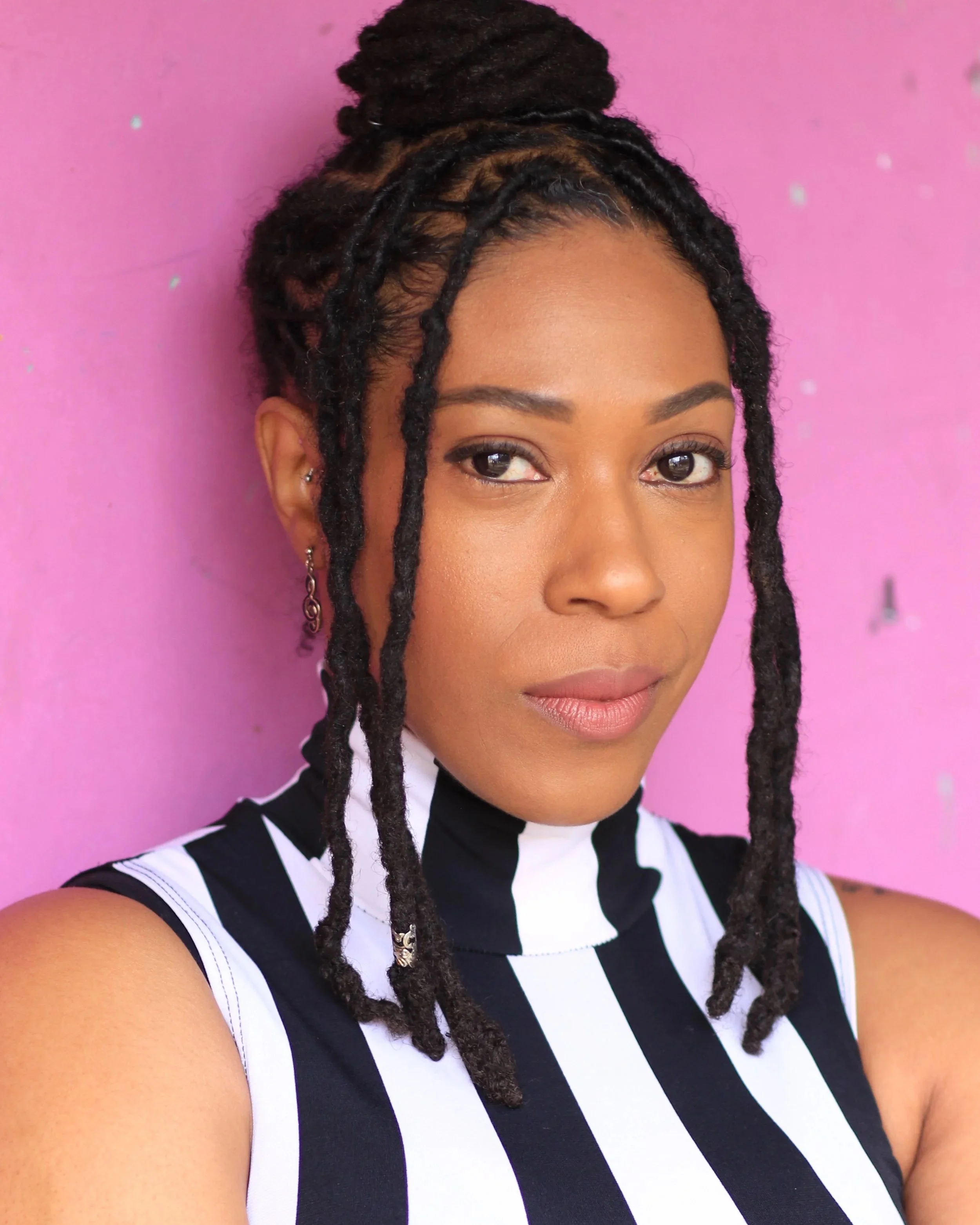 Close-up of an African American woman with styled dreadlocks in a bun, wearing earrings, against a pink background, all dressed in a black and white vertically striped top.