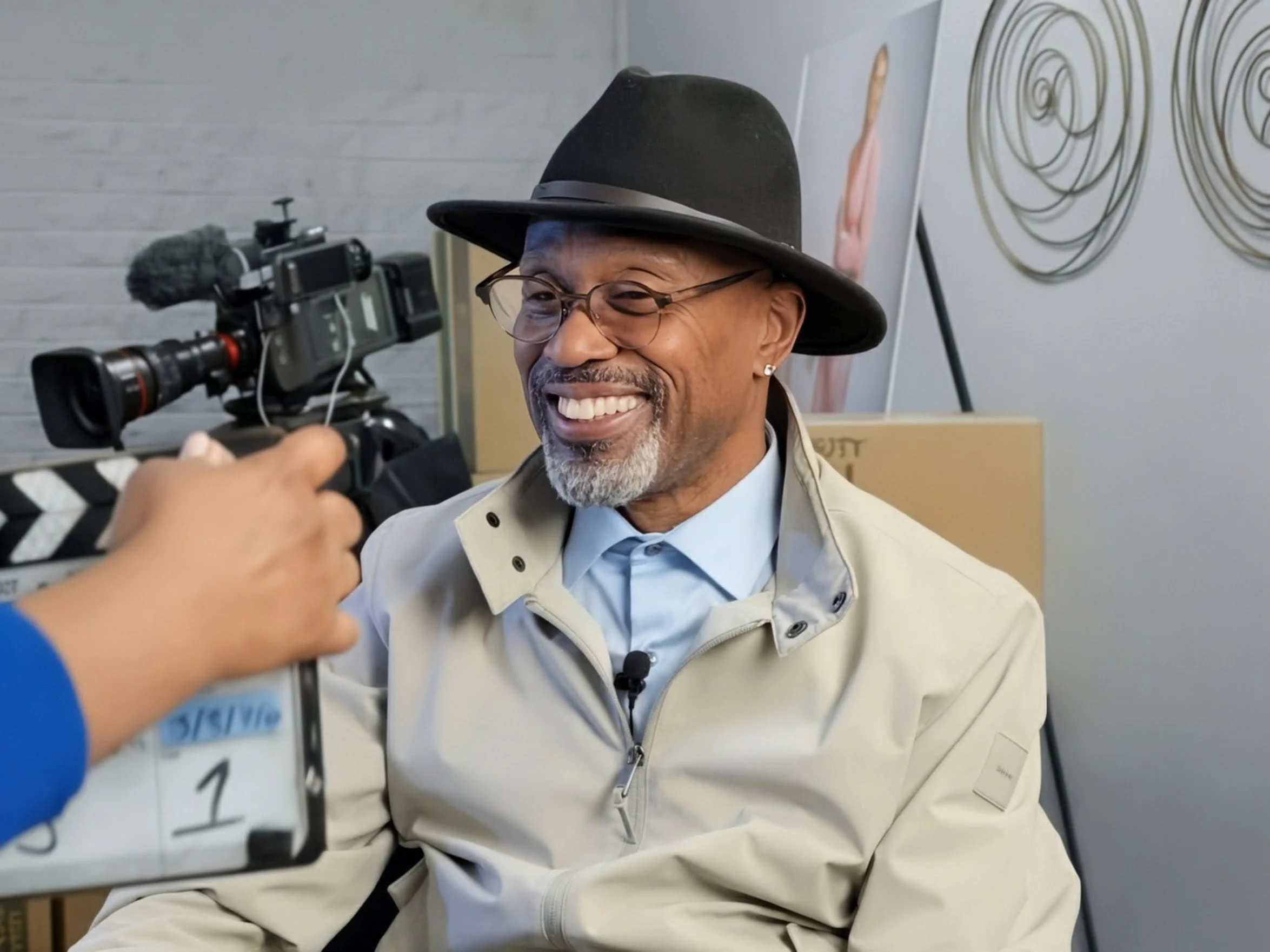 A smiling man wearing glasses, a black hat, a beige jacket, and a light blue shirt, being filmed with a professional camera in an indoor setting with white brick walls and abstract wall art in the background.
