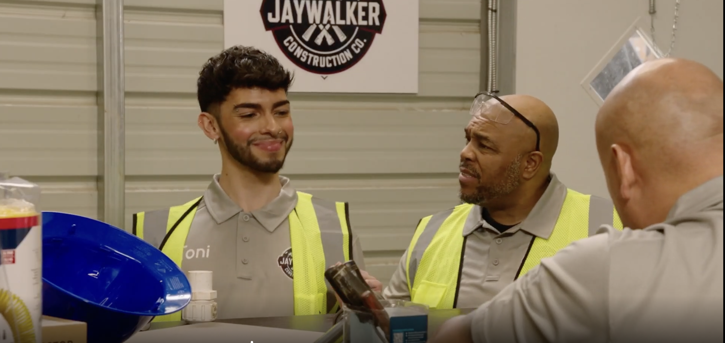 Three men wearing gray shirts and yellow safety vests having a conversation in a workshop or warehouse. There is a Jaywalker Construction Co. sign on the wall behind them, and various tools and supplies are visible on the counter.