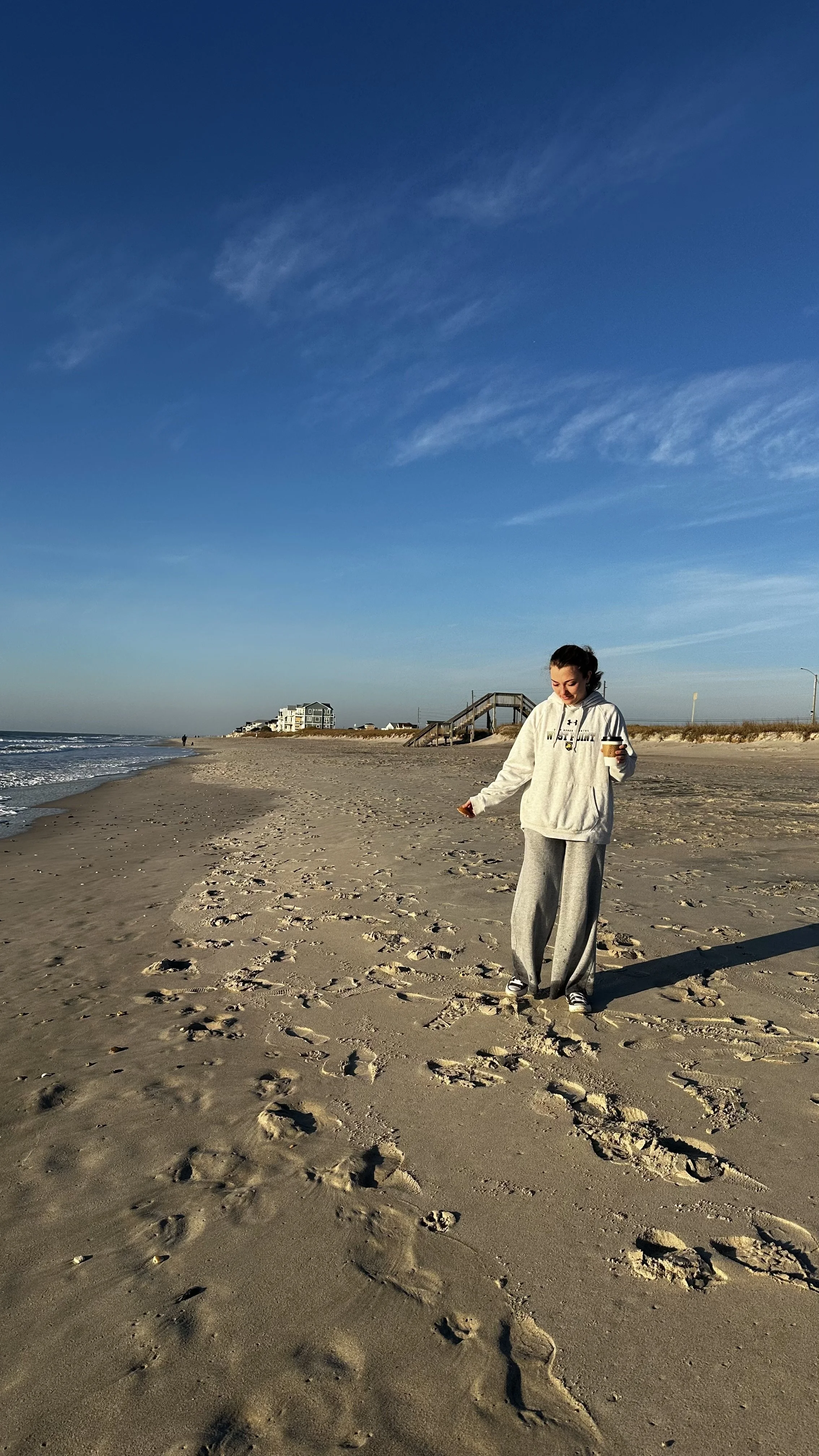 A woman walking barefoot on the sandy beach with footprints, holding a coffee cup, under a blue sky with some clouds, near ocean waves and beach houses in the background.