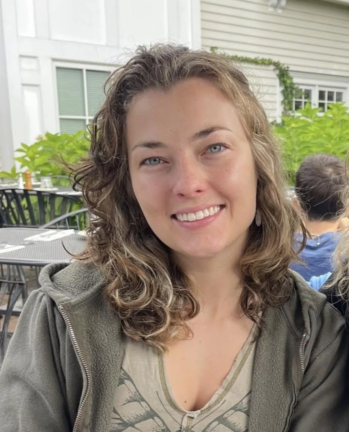 A woman with curly brown hair and blue eyes, smiling, seated outdoors at a restaurant with greenery and other diners in the background.