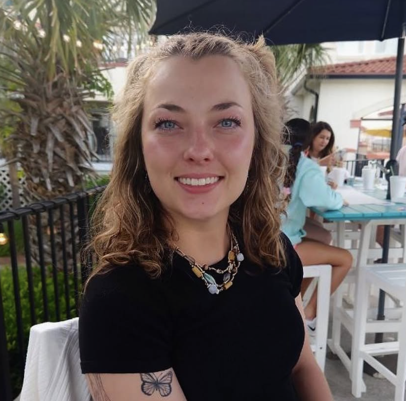 A smiling young woman with curly blonde hair and blue eyes sitting outdoors at a cafe or restaurant. She is wearing a black shirt and a beaded necklace. There are other people and tables in the background, along with umbrellas and greenery.