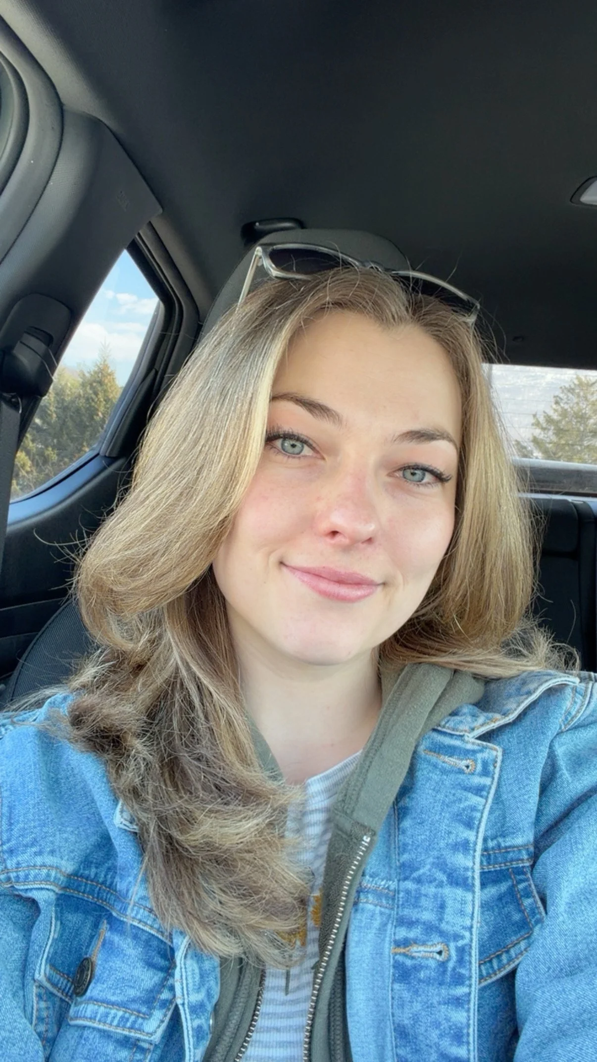 Young woman with light brown, wavy hair and blue eyes smiling in a car. She is wearing sunglasses on her head, a denim jacket, and a white and blue striped shirt, with a green hoodie underneath.