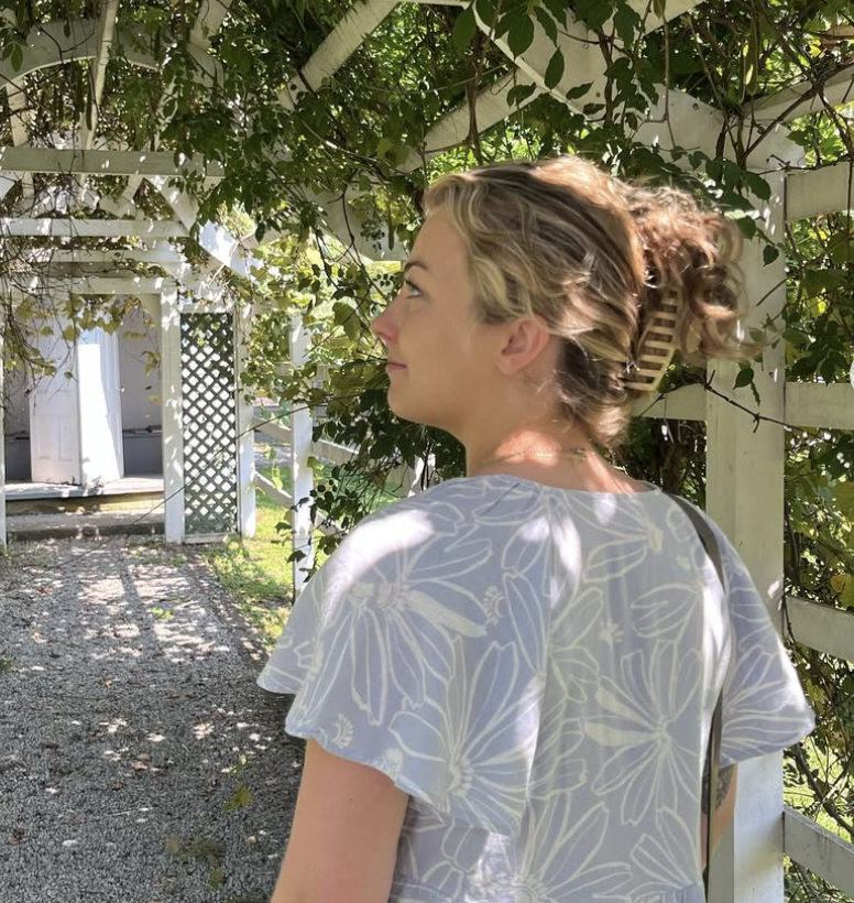 A woman with curly blonde hair and a wooden hair clip, wearing a white dress with a floral pattern, standing with her profile visible against a background of green leaves and a white wooden fence.