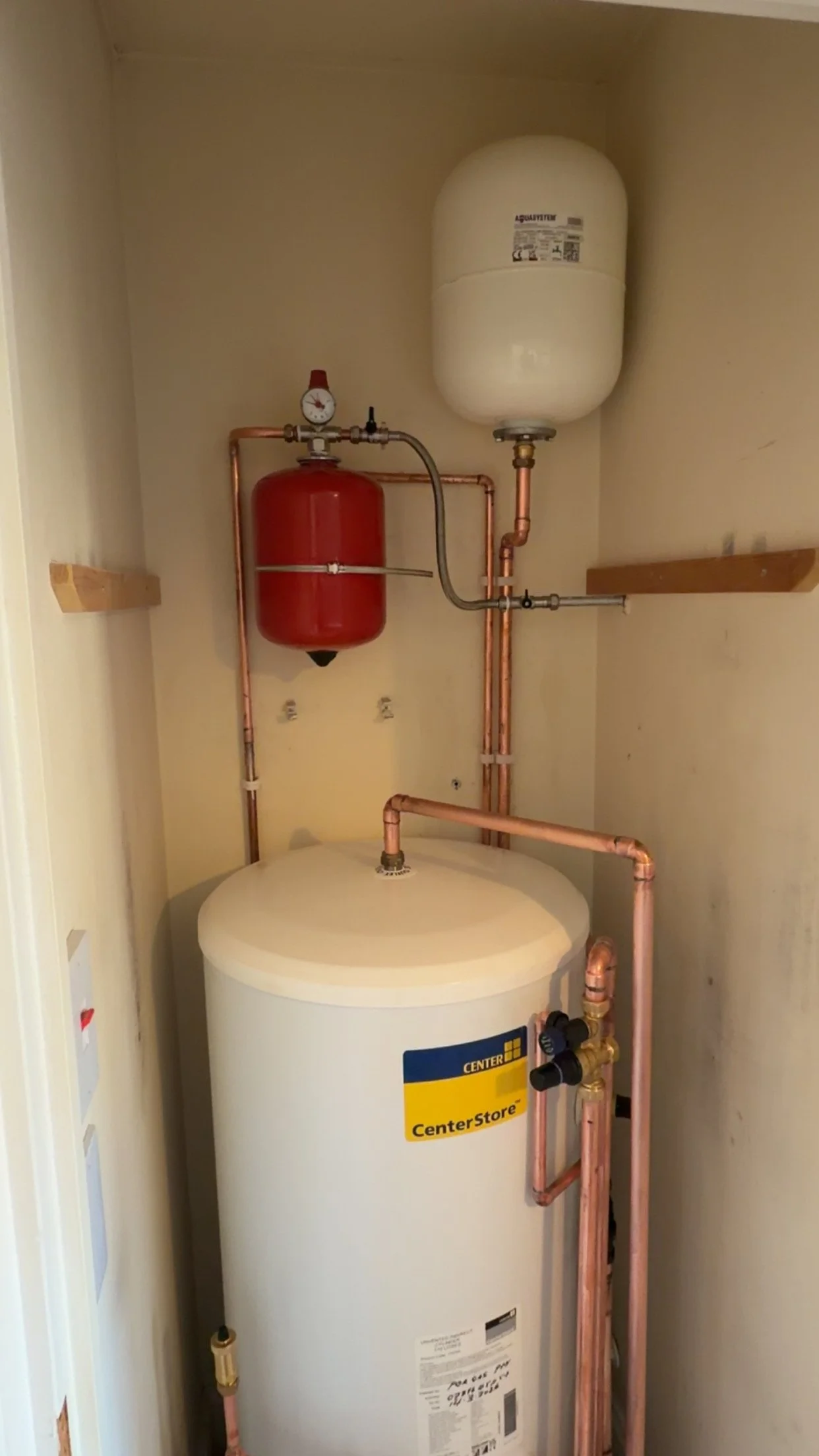 A water heater unit with copper pipes, a red expansion tank, and an expansion tank with a pressure gauge in a small utility closet.