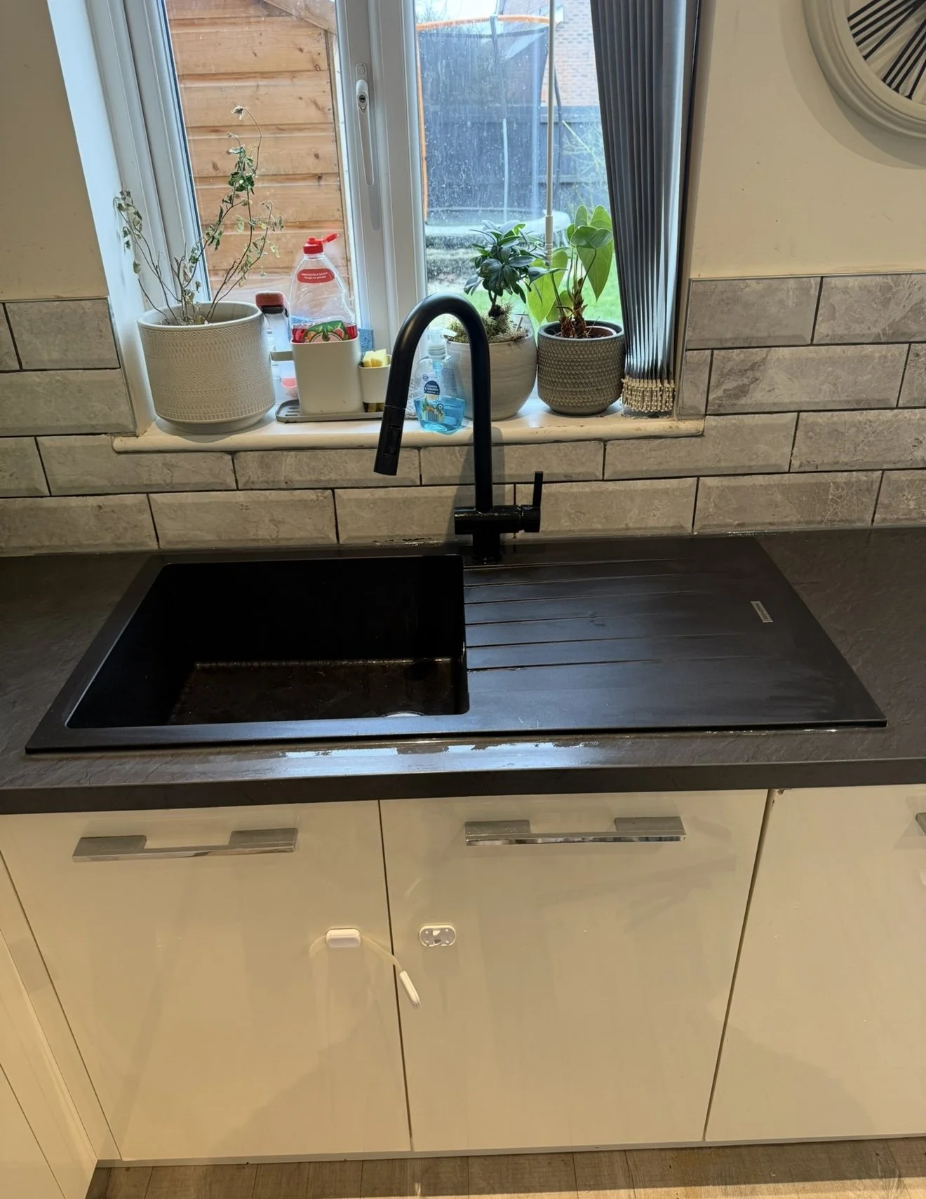 A kitchen sink with a black faucet, white cabinet with silver handles, a window with three potted plants on the windowsill, a bottle of hand soap, and various cleaning supplies on the window ledge.