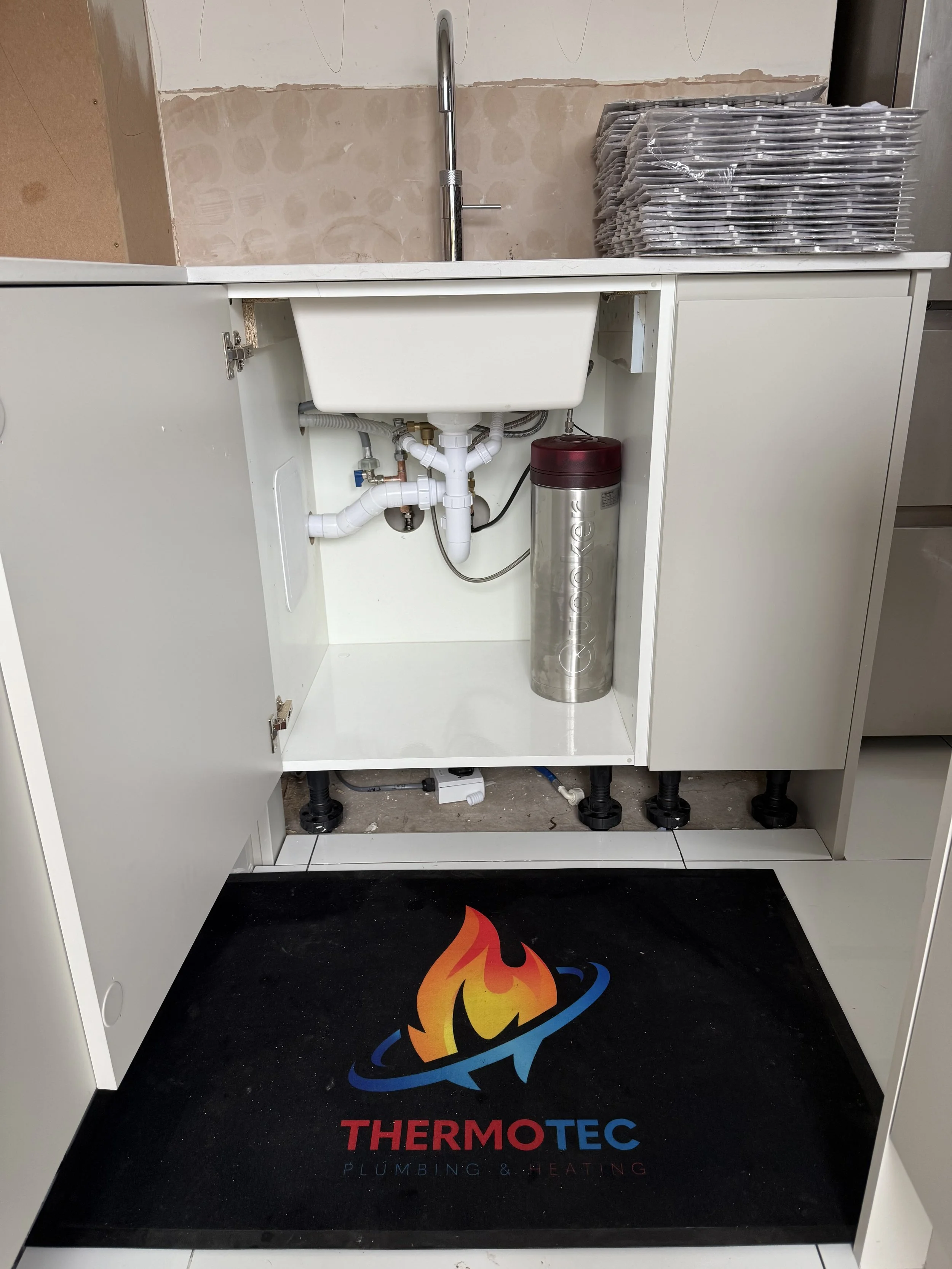 Underneath a kitchen sink cabinet, plumbing pipes are visible, with a water filter attached. To the right, there are stacked trays or containers. The floor has a black mat with a colorful flame logo and the words 'THERMOTEC Plumbing & Heating'.