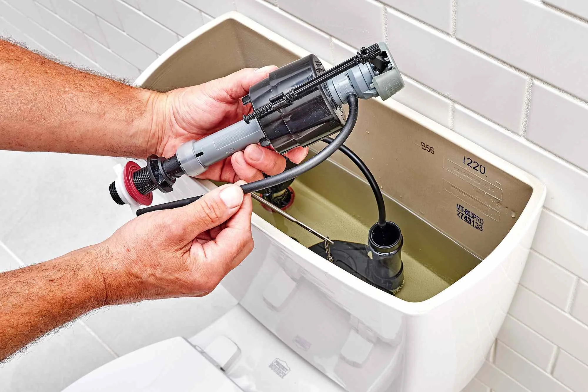 A person holding a toilet fill valve above a toilet tank, about to install or replace it.