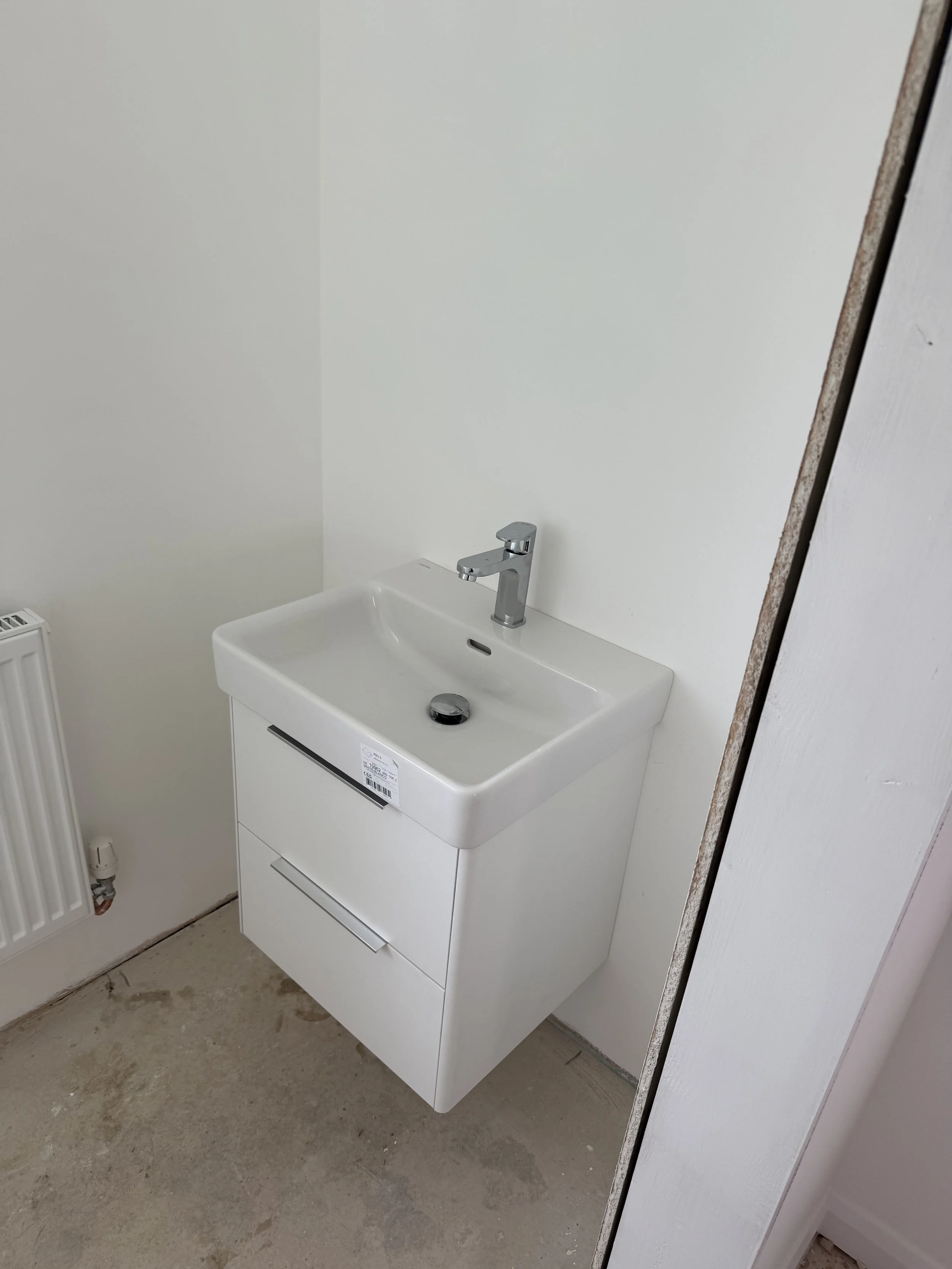 A white bathroom vanity with a sink and a chrome faucet, mounted against a plain light-colored wall, with a radiator and some flooring visible.