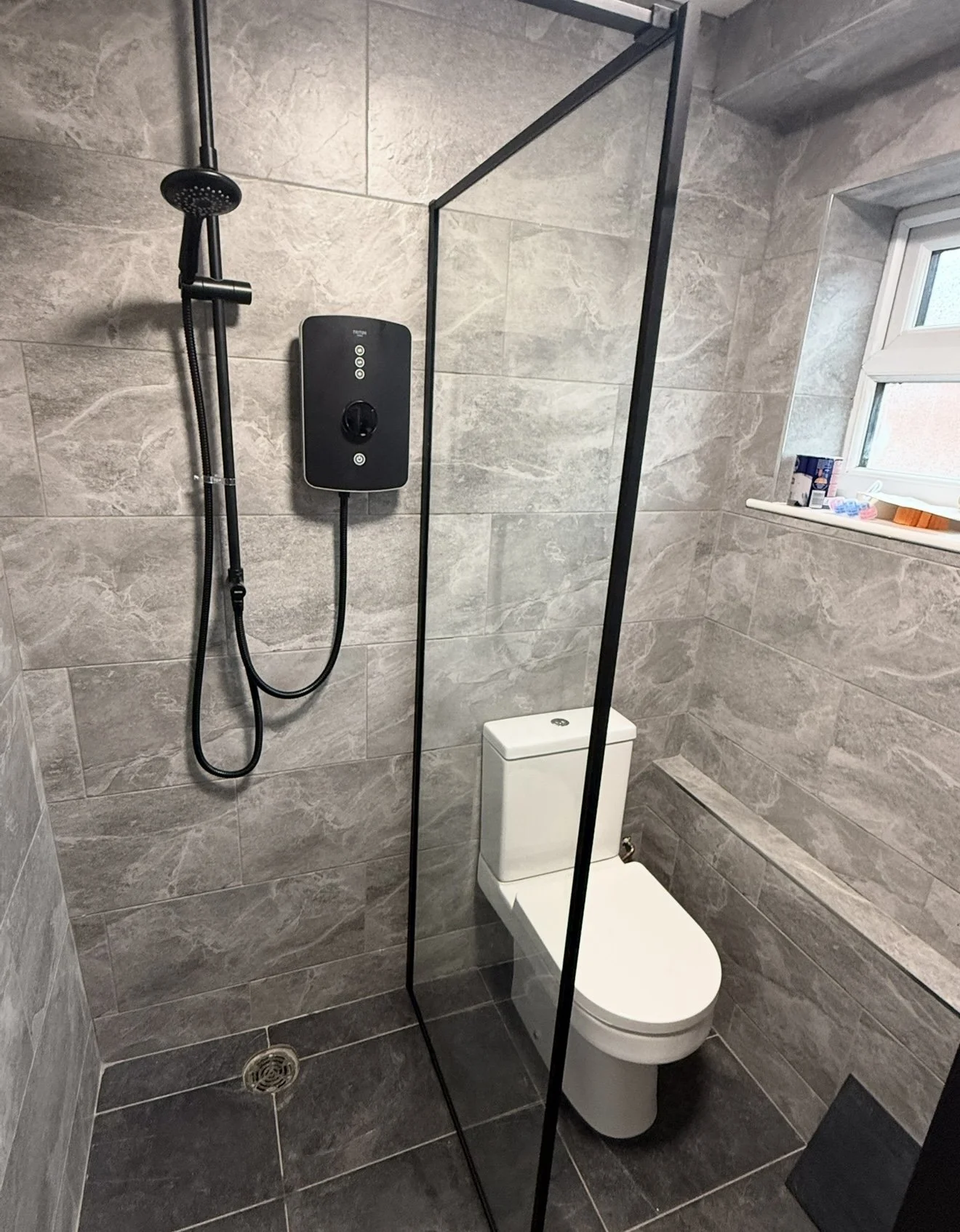 Modern bathroom with gray marble tiles, a toilet, a black shower panel with a handheld showerhead, a glass divider, and a small window on the right with a windowsill holding toiletries.