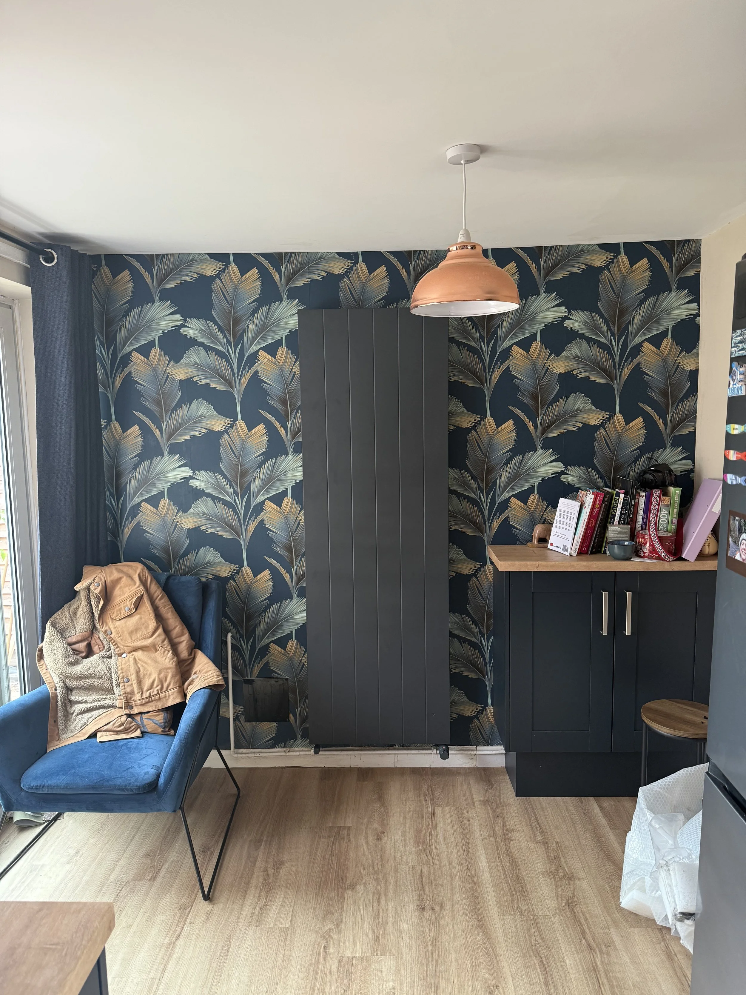 Interior of a room with dark blue wallpaper with a tropical leaf pattern, a black cabinet, a blue velvet chair with a tan jacket on it, a small wooden table, and a window with gray curtains. A copper-colored hanging light fixture is above, and a book