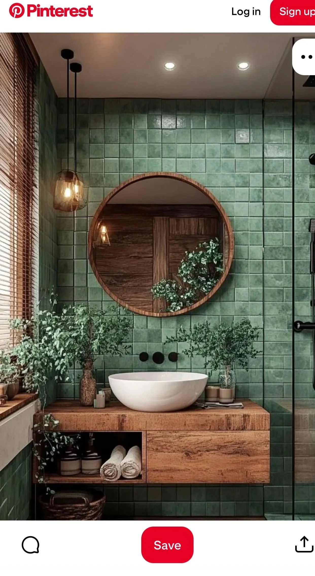 A modern bathroom with green tiled walls, a white round bathtub on a wooden platform, a large circular mirror, green plants, and wooden accents. There are pendant lights hanging from the ceiling and a window with bamboo blinds.