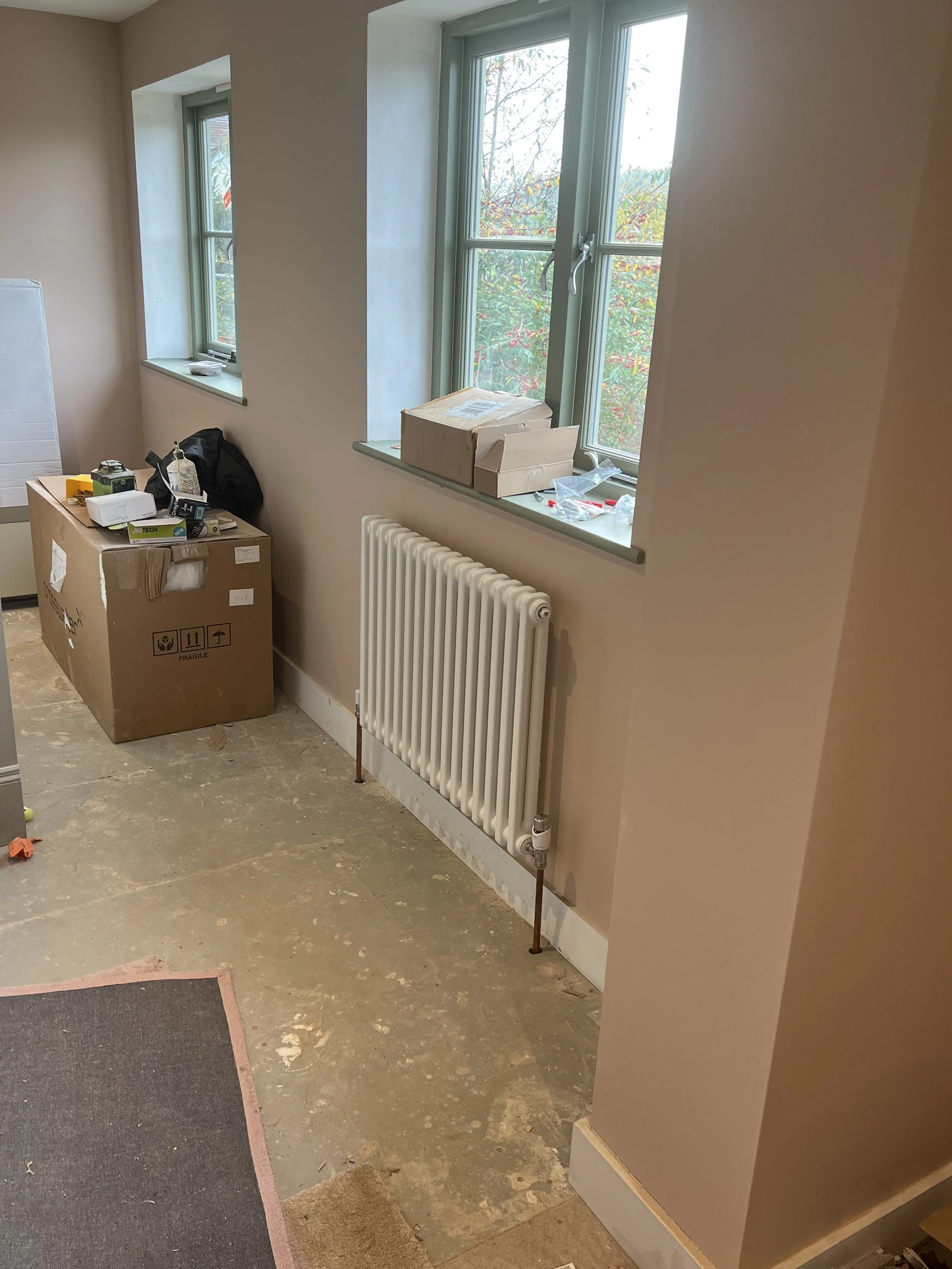 Interior of a room under renovation with a radiator, window sills with boxes and tools, and a partially finished floor.