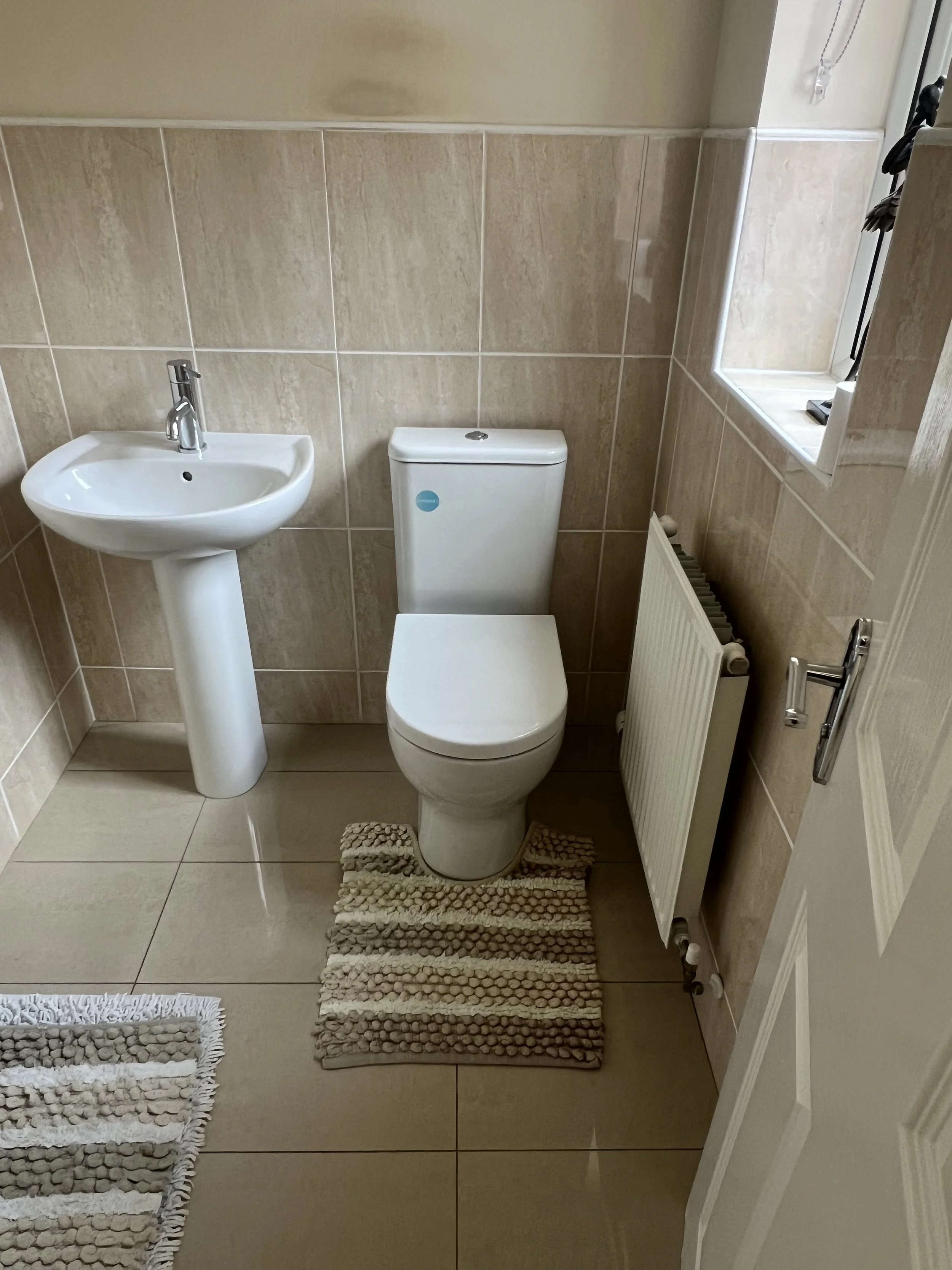 Small bathroom with beige tiled walls and floor, a white toilet and a pedestal sink, a radiator, a window with a lace curtain, and a beige rug in front of the toilet.
