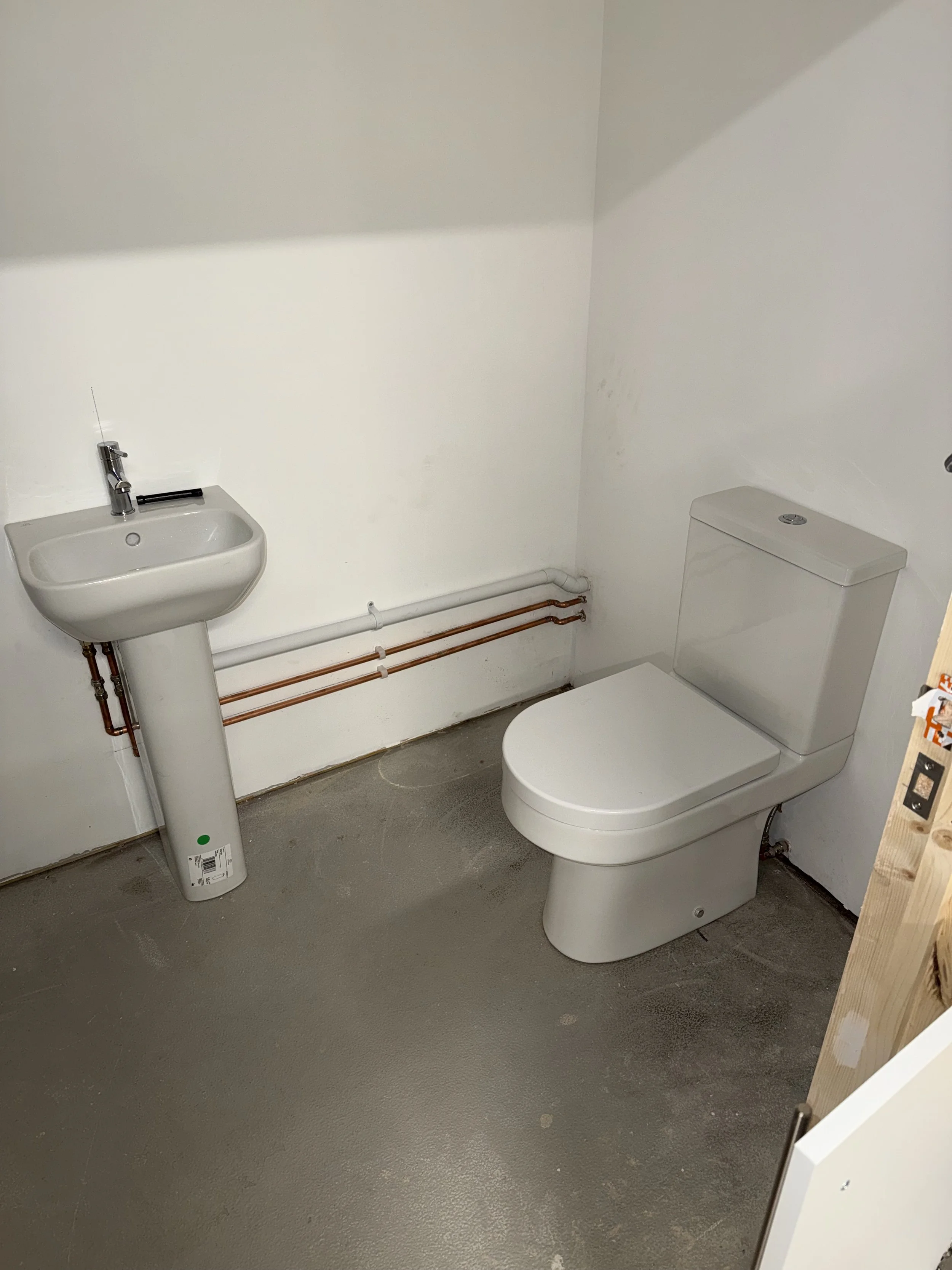 A small bathroom with a white toilet and a pedestal sink. Visible pipes run along the wall under the sink, and the floor appears to be concrete.