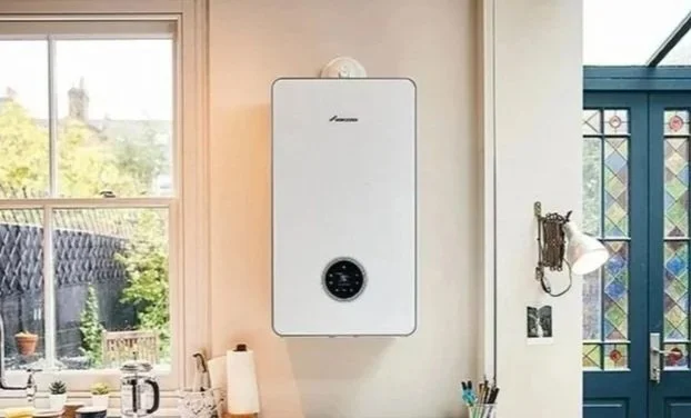 A white wall-mounted boiler in a kitchen with a window showing greenery outside, a paper towel roll, and kitchen utensils on the countertop.