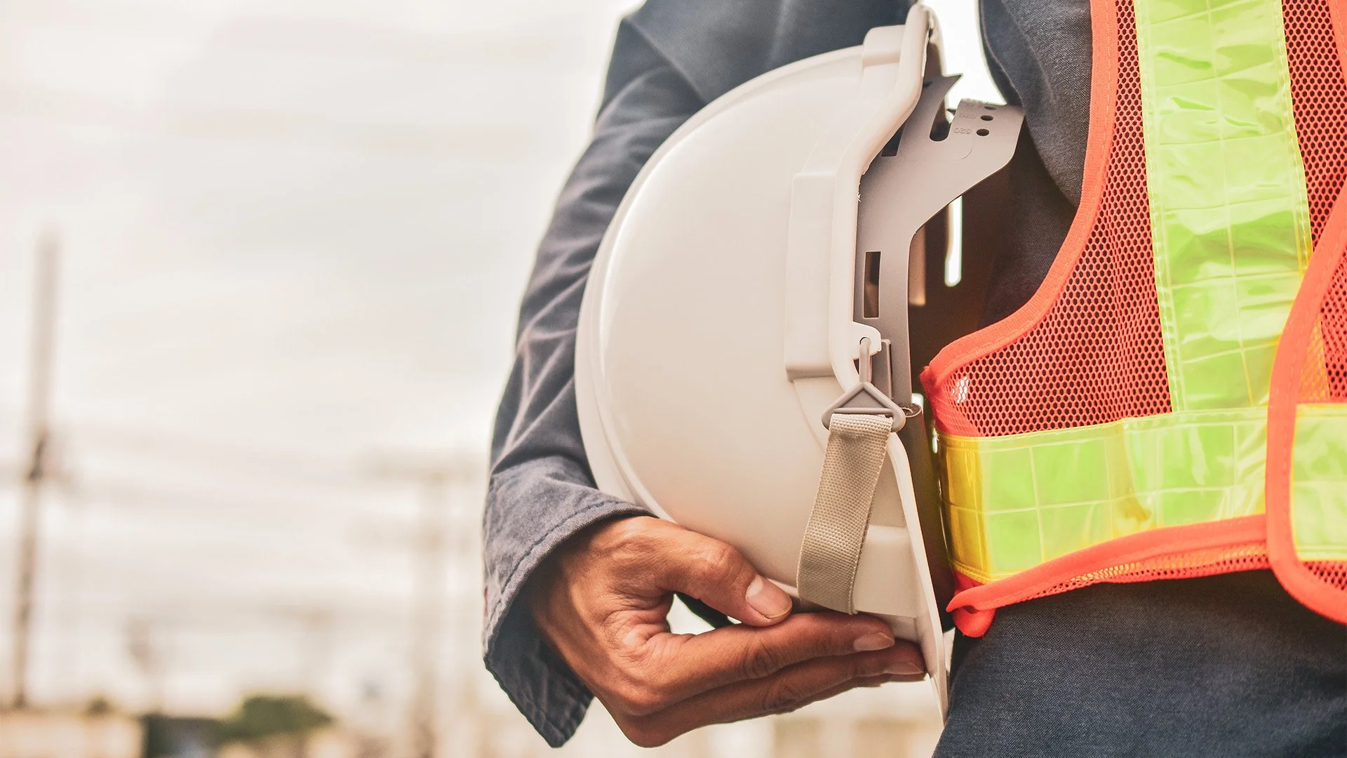 A person holding a white safety helmet with a strap, wearing a neon safety vest with orange trim, in an outdoor construction site.
