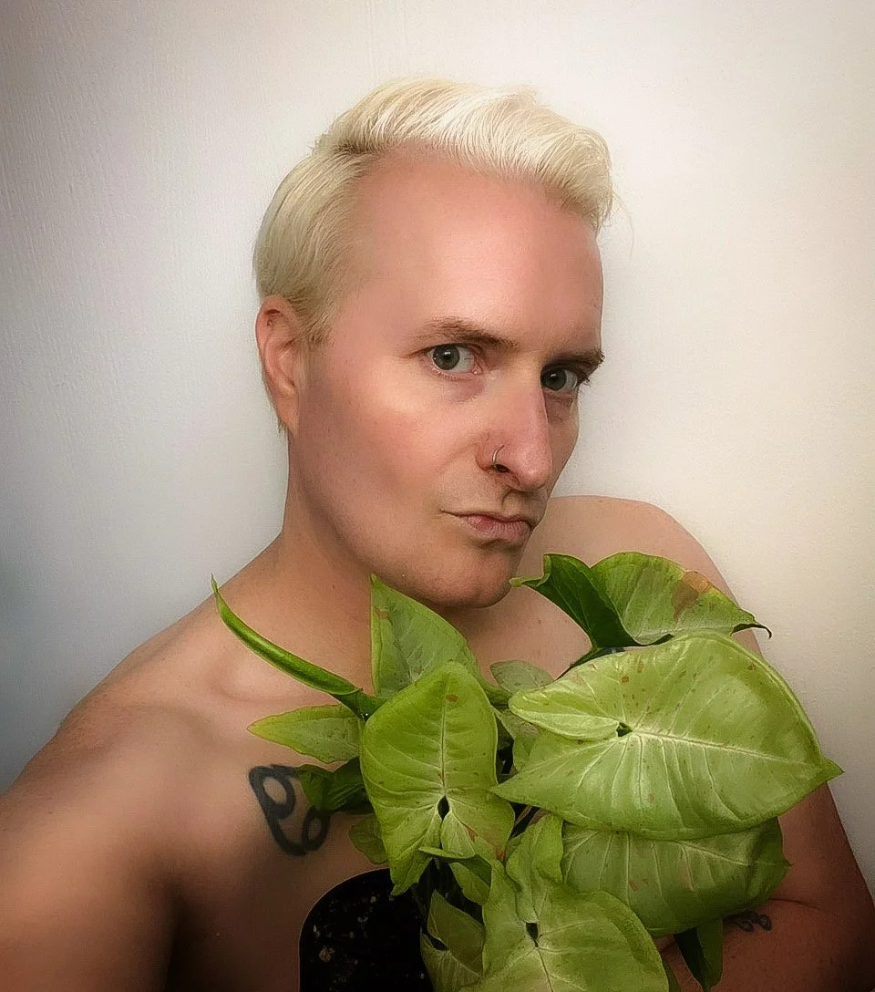A person with bleach blonde hair and a nose ring posing with a large green leafy plant. The person is shirtless and has a tattoo on their upper arm.