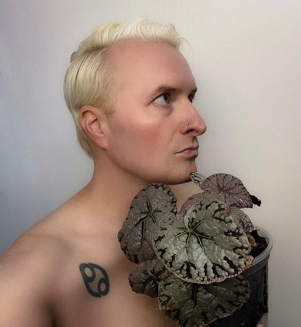 Person with short blonde hair and a nose ring holds large purple and green patterned leaves close to their shoulder with a plain light-colored wall in the background.