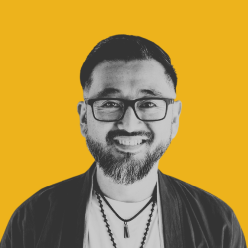 Smiling person with dark hair, dark frame glasses, a trim beard, and necklaces.