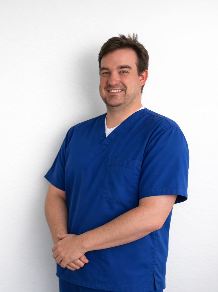 A male healthcare professional in blue scrubs standing against a white wall, smiling and looking at the camera.
