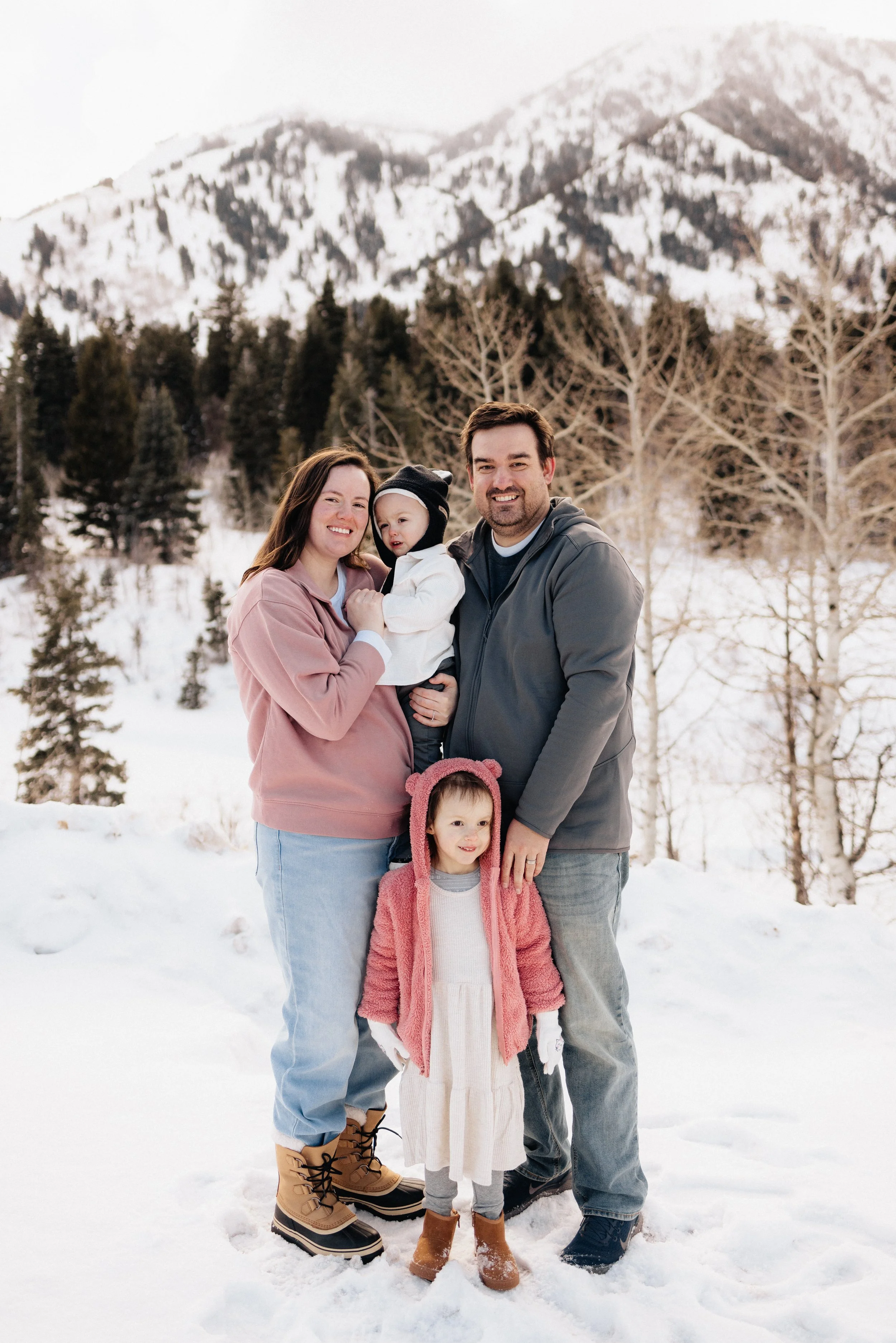 Family of four standing in snowy landscape with mountains and trees in the background, dressed in winter clothing.