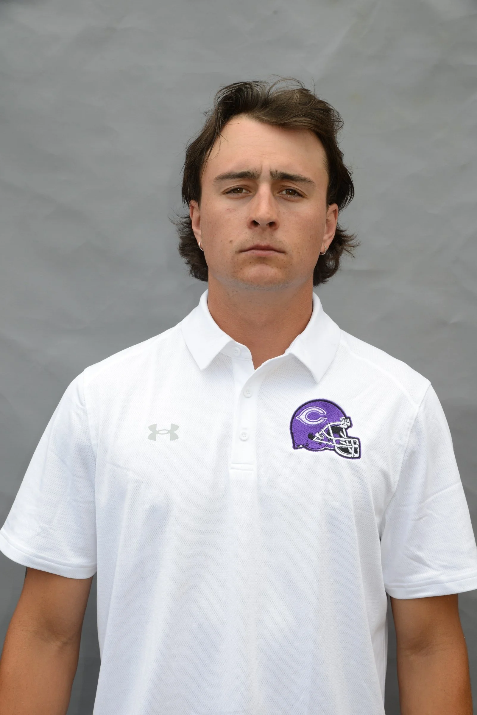 A young man with dark hair, wearing a white polo shirt with a purple and black football helmet logo on the chest, standing against a gray cloudy sky.