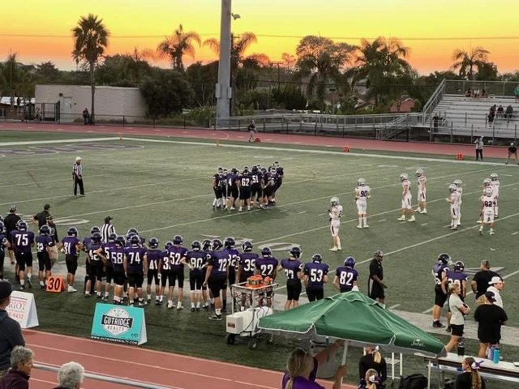 Players on the football field during a game at sunset, with team huddles and spectators in the stands.
