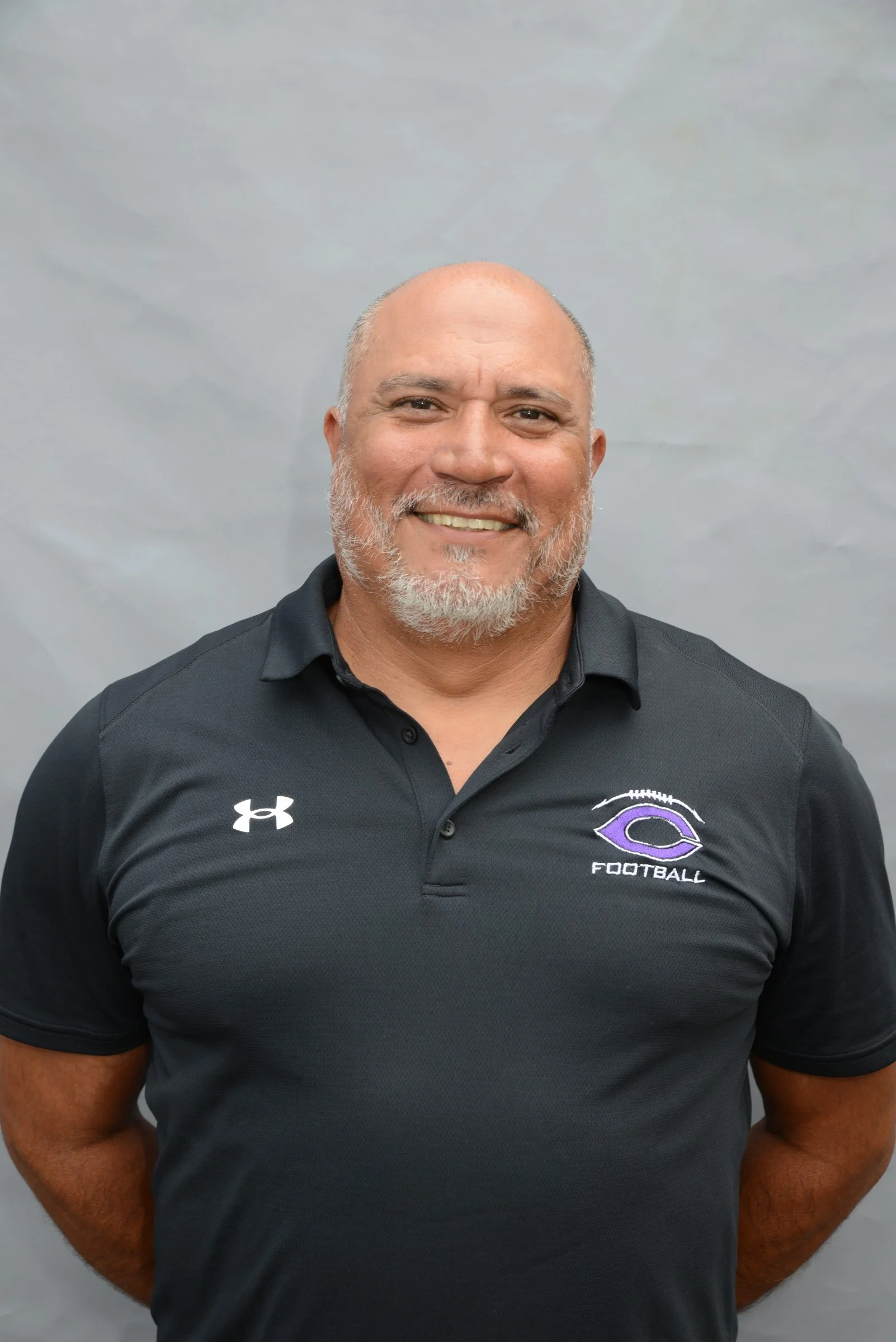 A smiling man with a beard and shaved head standing against a plain background. He is wearing a black Under Armour polo shirt with a purple football logo and the word 'FOOTBALL' on it.
