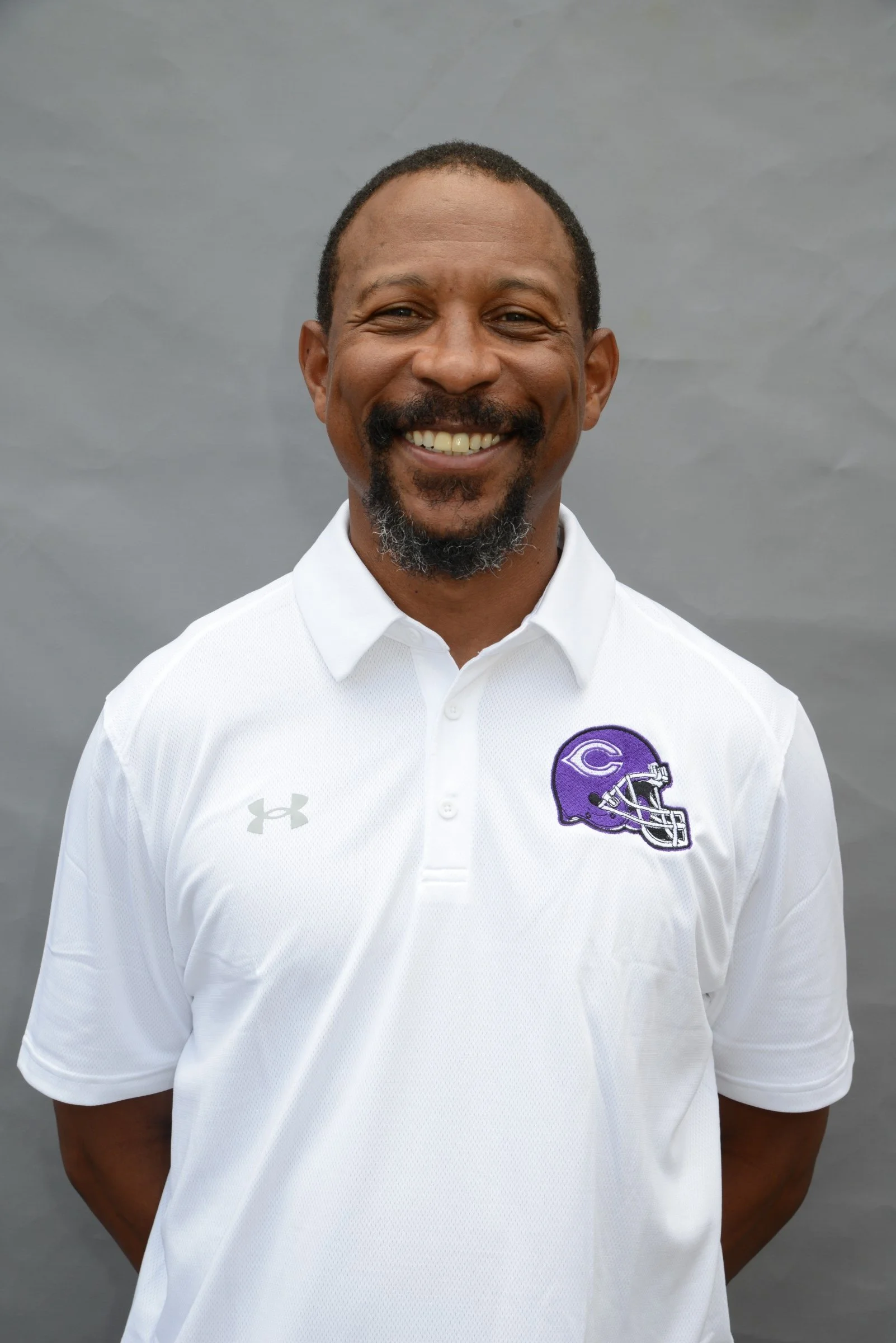Smiling African American man wearing a white polo shirt with a purple football helmet logo on the chest, standing against a gray background.