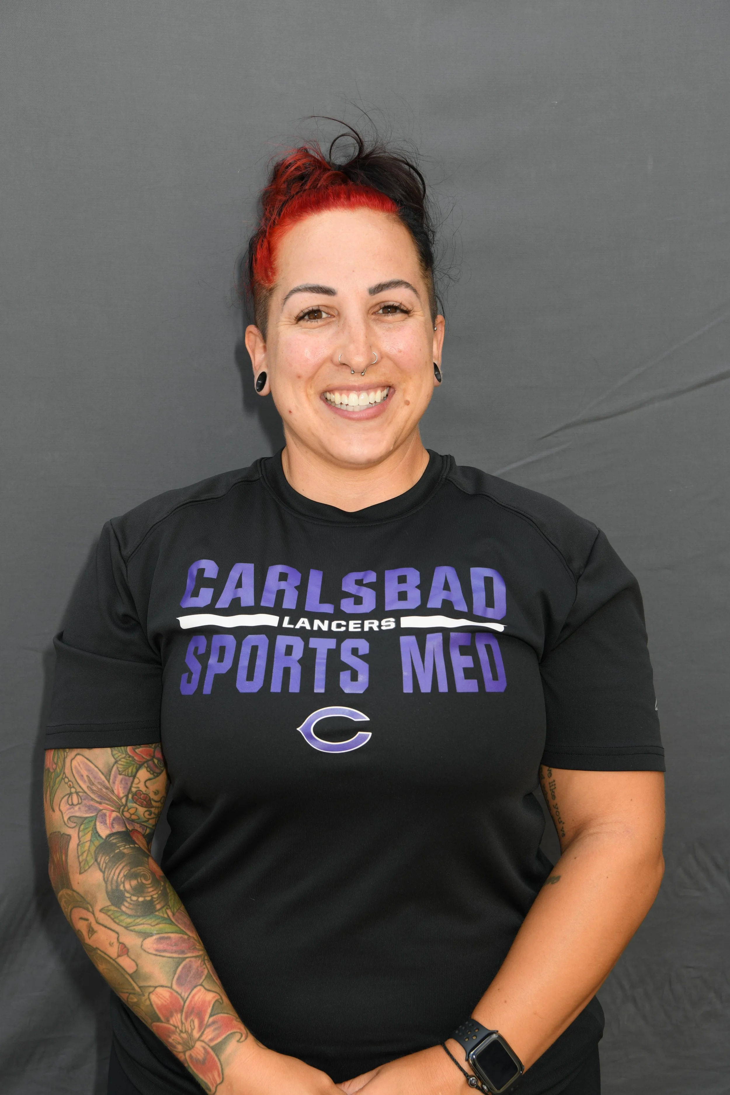 A smiling woman with dark and red hair in a bun, wearing a black sports shirt with purple and white text, standing against a gray background. She has tattoos on her left arm, black earrings, a nose ring, and a smartwatch on her left wrist.