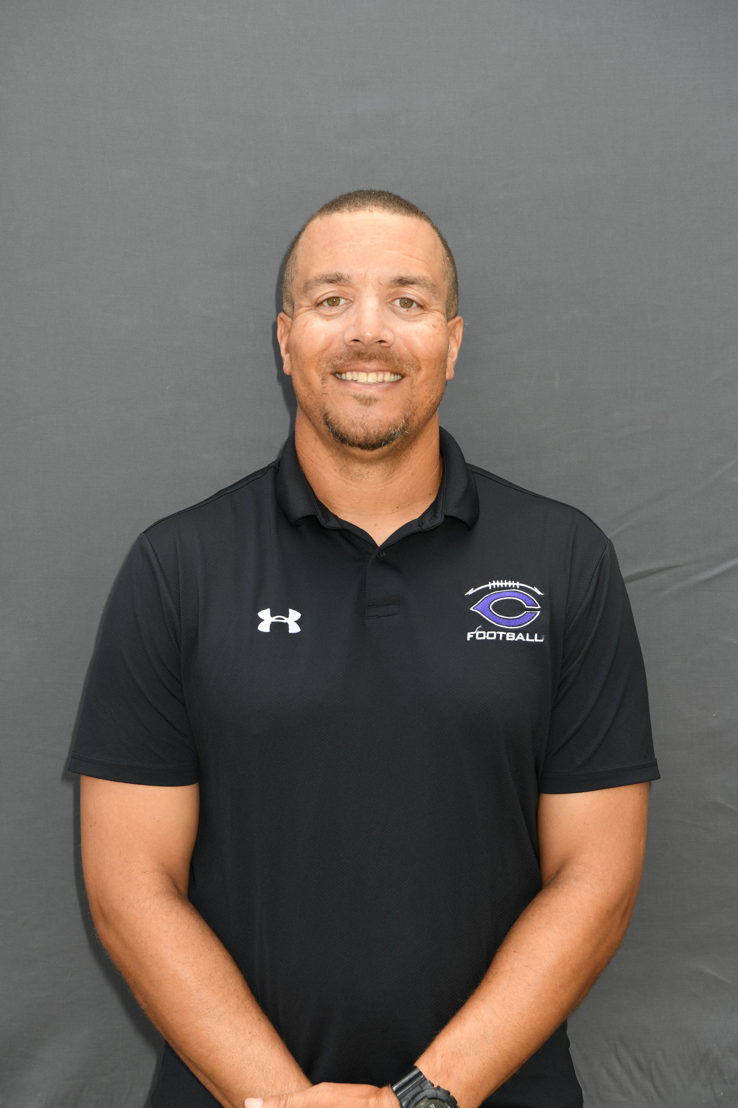 Portrait of a smiling man in a black polo shirt with a football team logo, standing in front of a gray background.