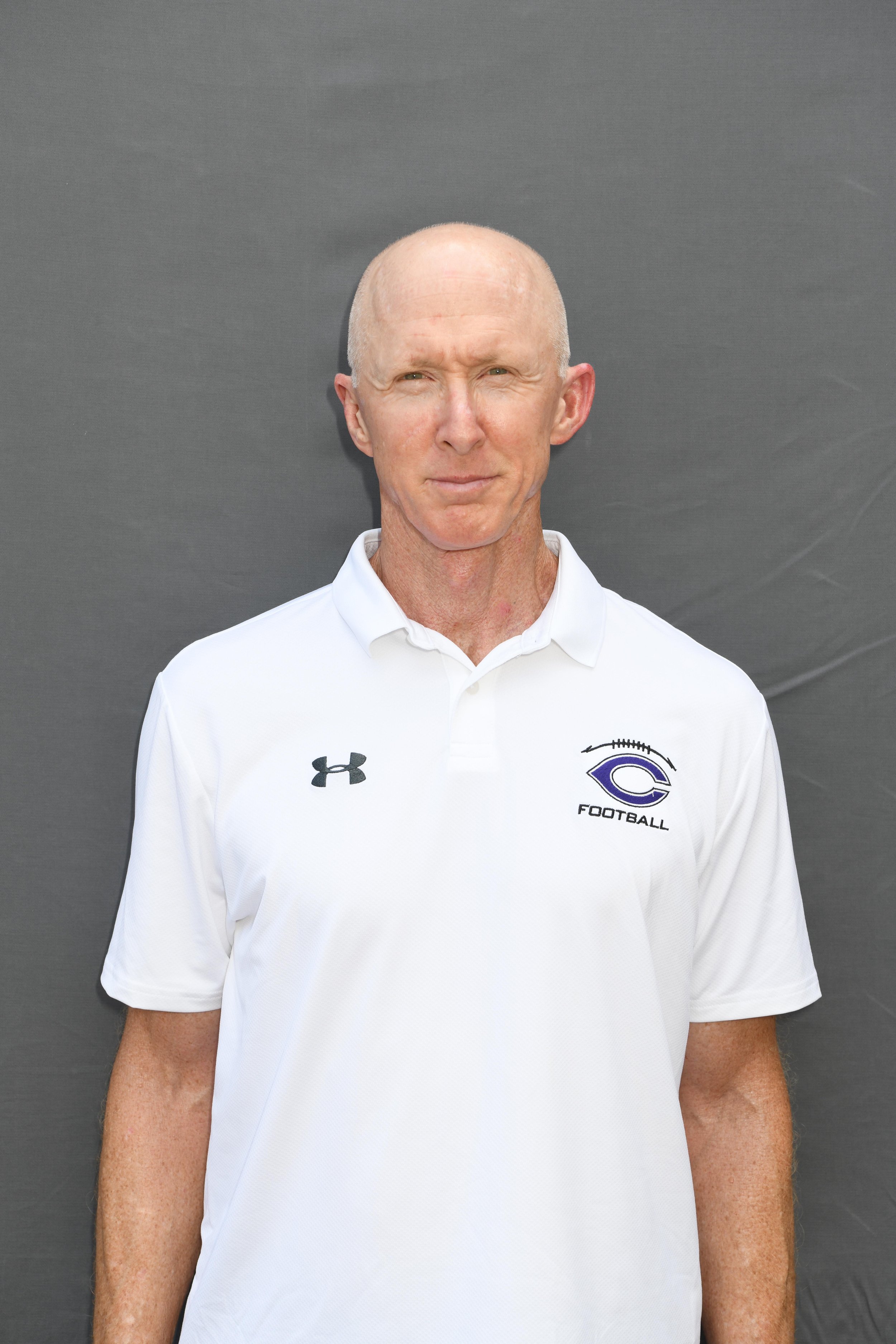 A bald man in a white sports polo shirt with Under Armour logo and a football team logo, standing against a gray background.