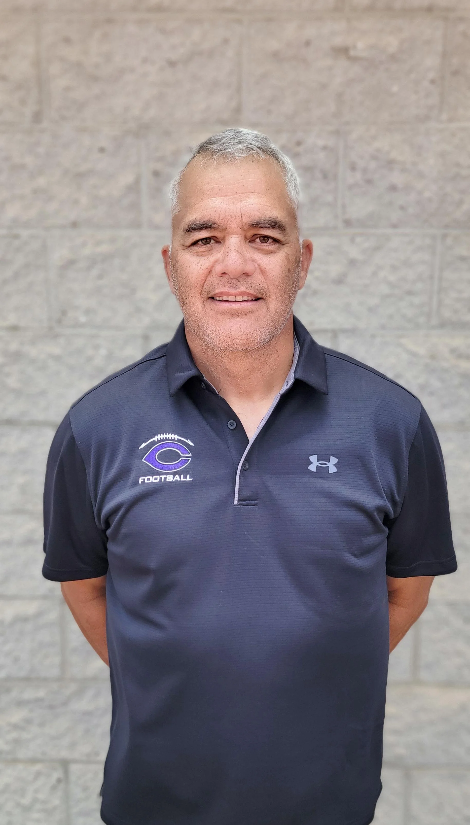A middle-aged man with short gray hair and stubble, smiling, standing in front of a light gray brick wall. He is wearing a black Under Armour polo shirt with football team logos on the chest.