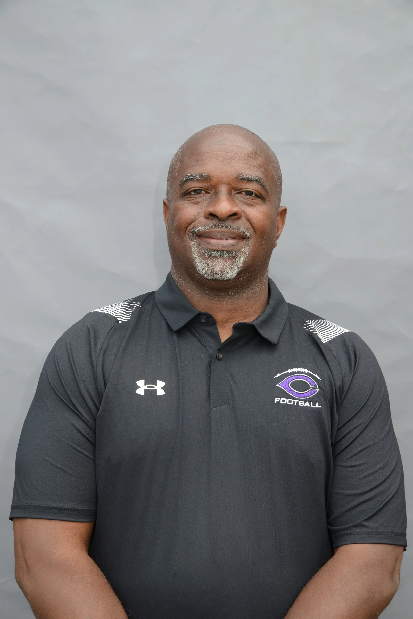 A smiling man wearing a black football coach shirt with a logo, standing against a gray background.