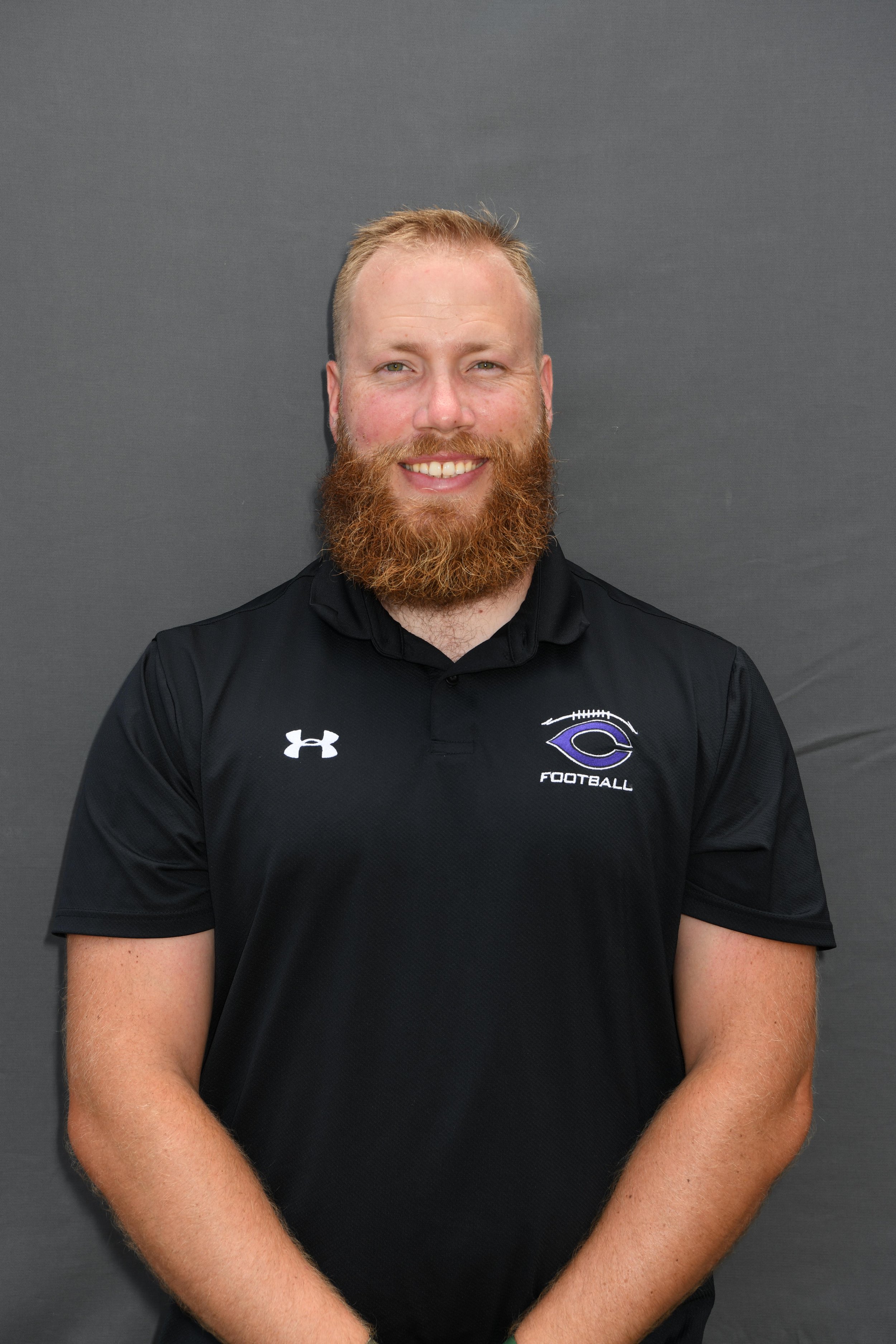 A man with a red beard and short light hair, wearing a black sports shirt with a team logo, smiling, standing against a gray background.