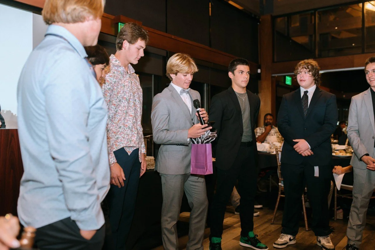 Group of young men in formal attire at an indoor event, one speaking into a microphone, holding a purple gift bag, with people seated at tables in the background.