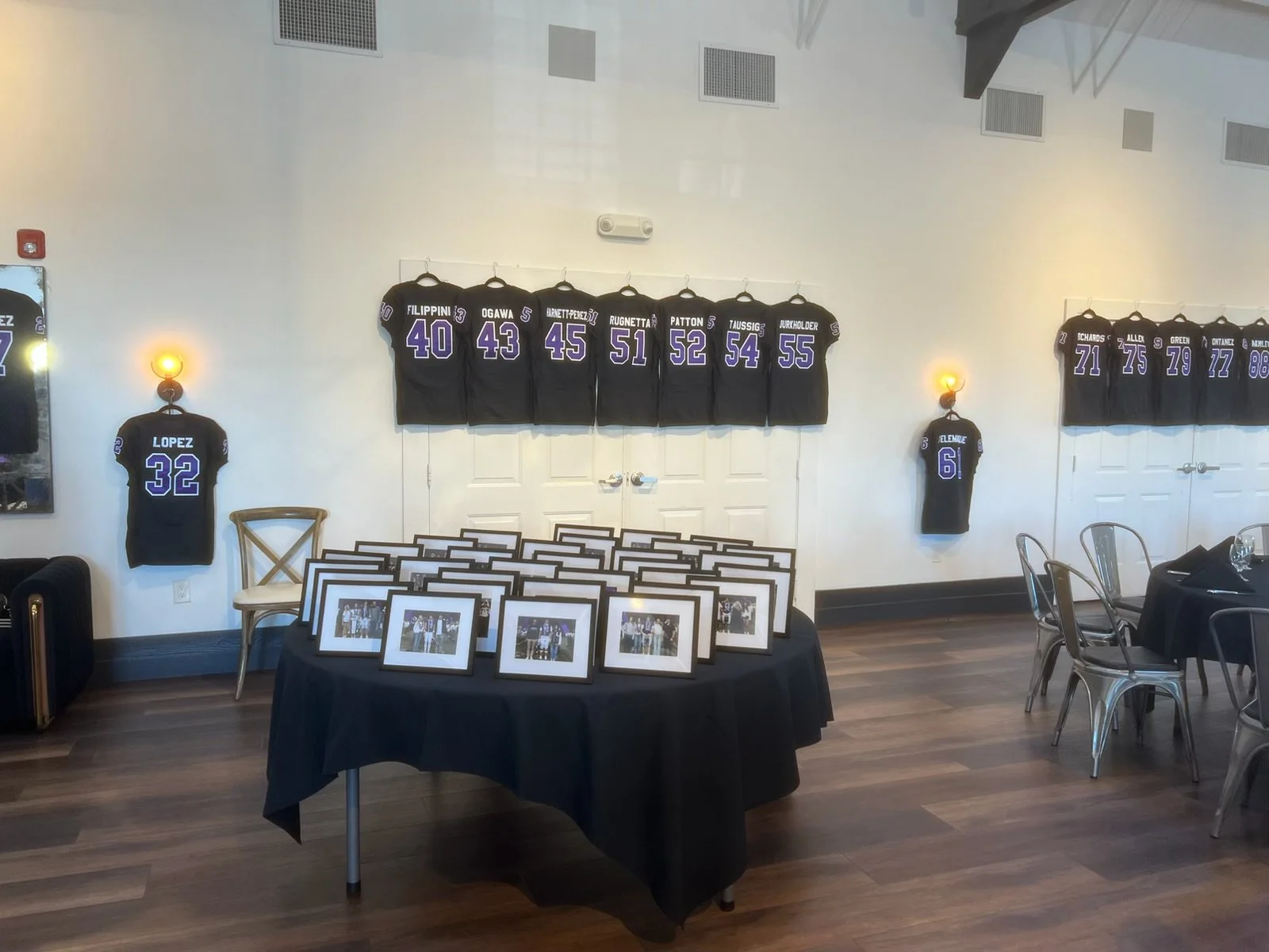 Display of black jerseys with white and purple numbering on a wall, framed photographs on a table in the foreground, and chairs around a table in a room with wooden flooring.