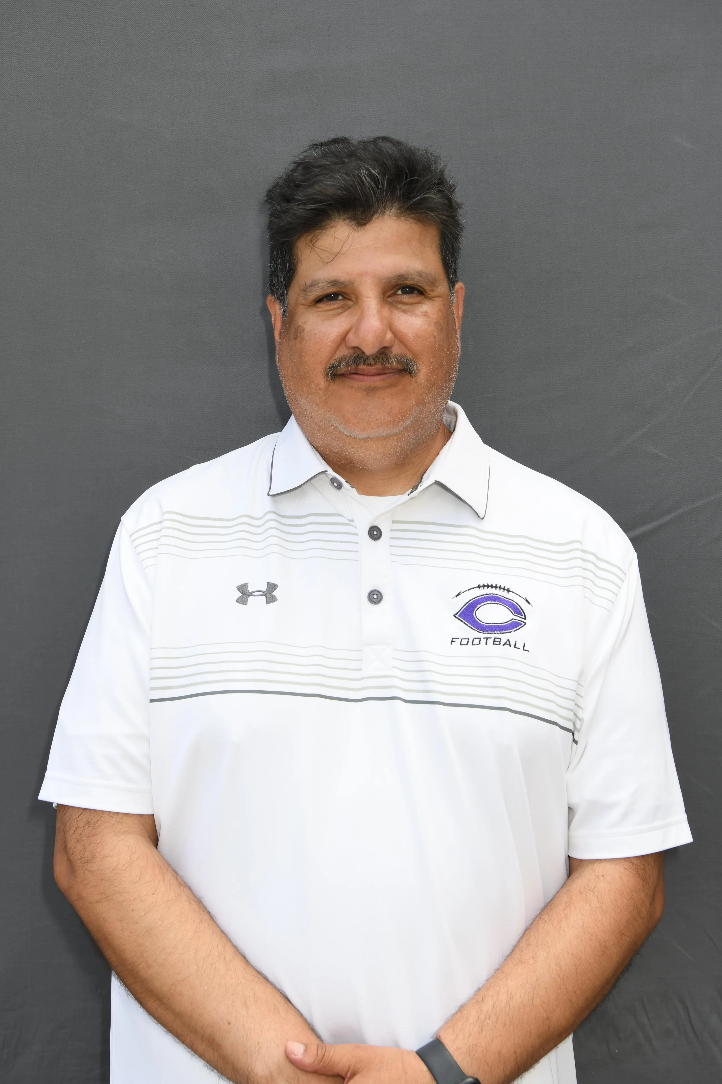 A man in a white football team shirt with a logo that says 'football' and a purple stylized 'C' logo on it, standing against a gray background, with arms crossed.