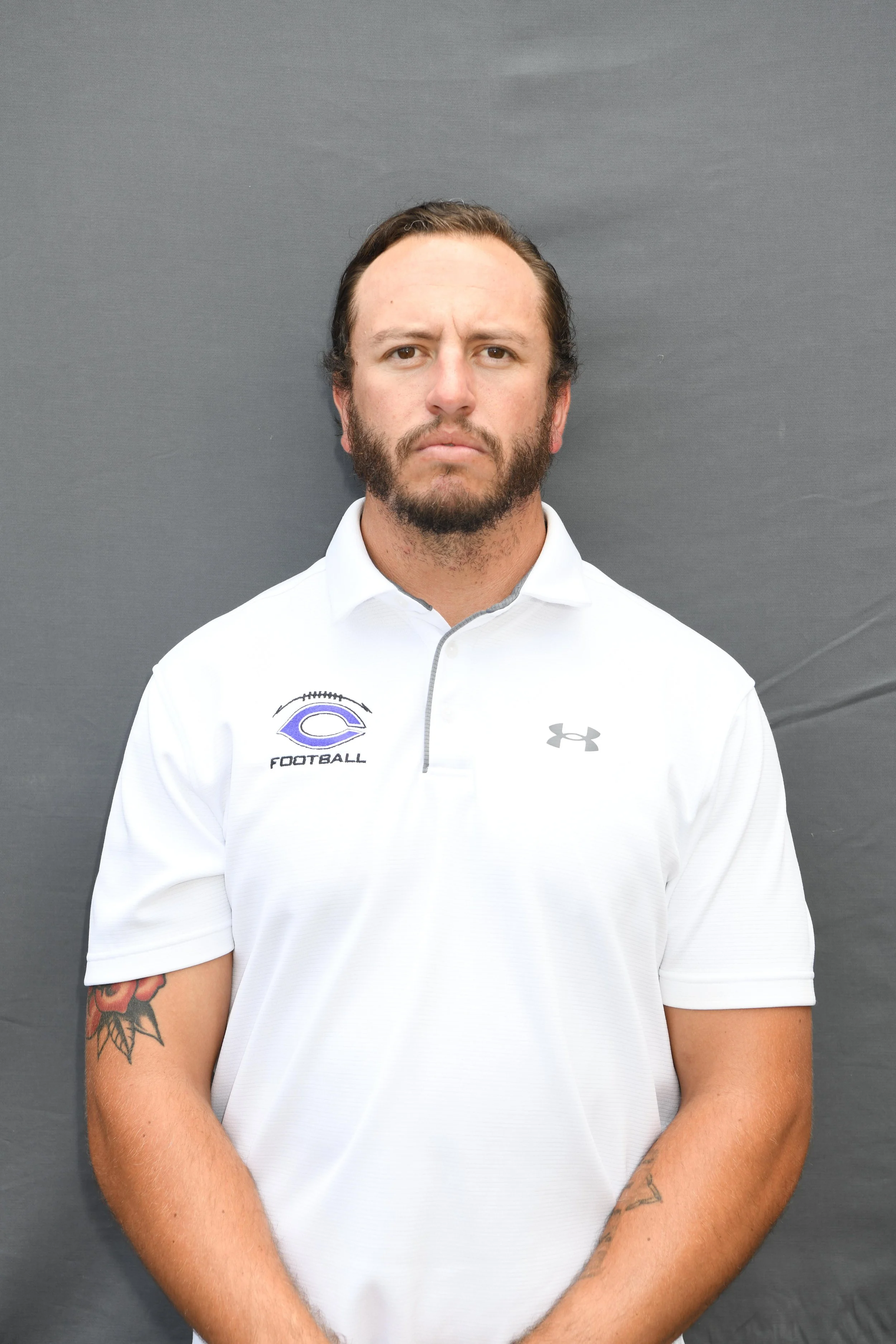 A man with dark hair and a beard stands against a plain dark grey background, wearing a white sports shirt with logos on the chest and an Under Armour logo. He has tattoos on his right forearm.