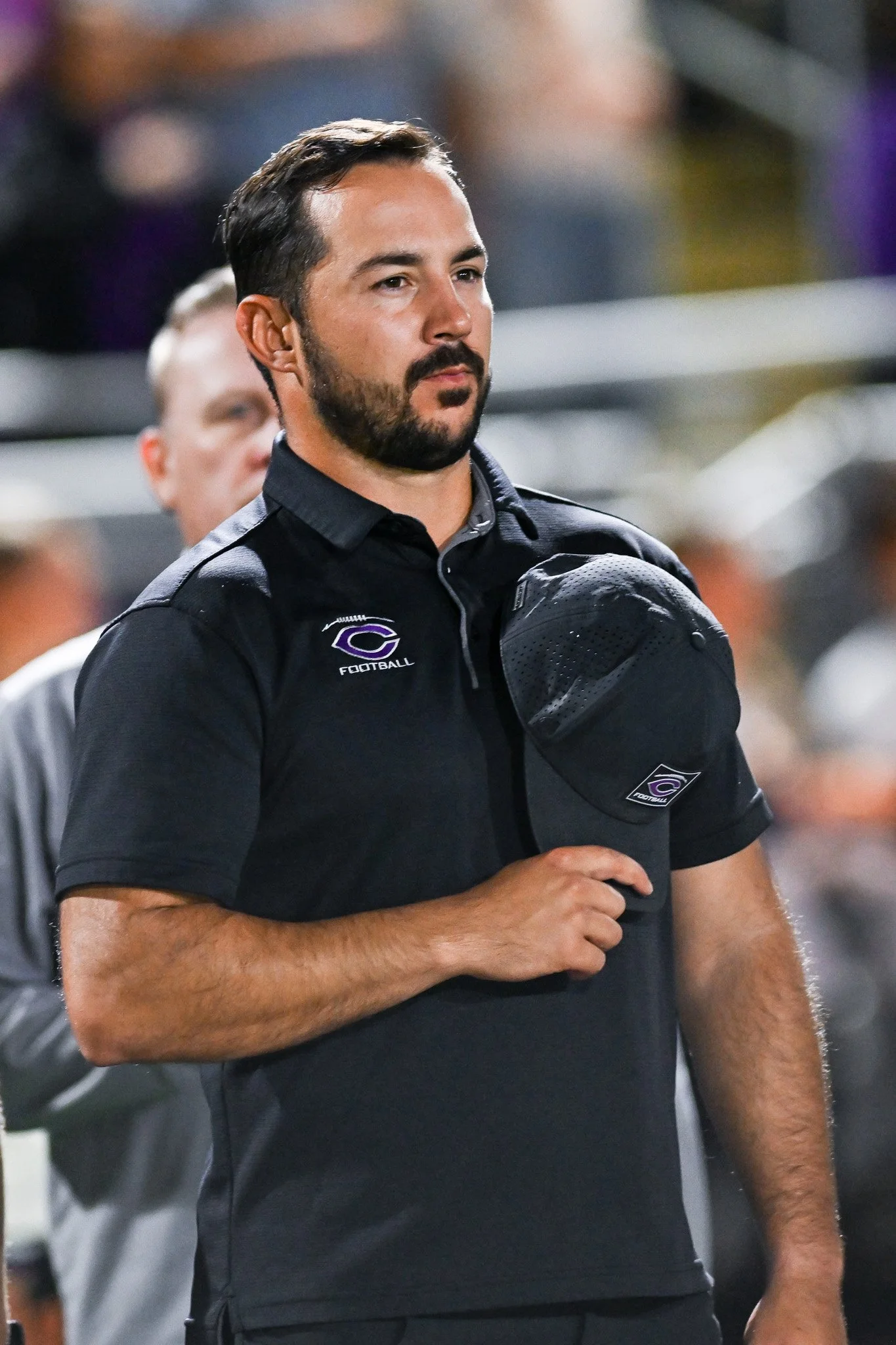 A man with dark hair and a beard, wearing a black shirt with a football team logo, standing outdoors with blurred people in the background.