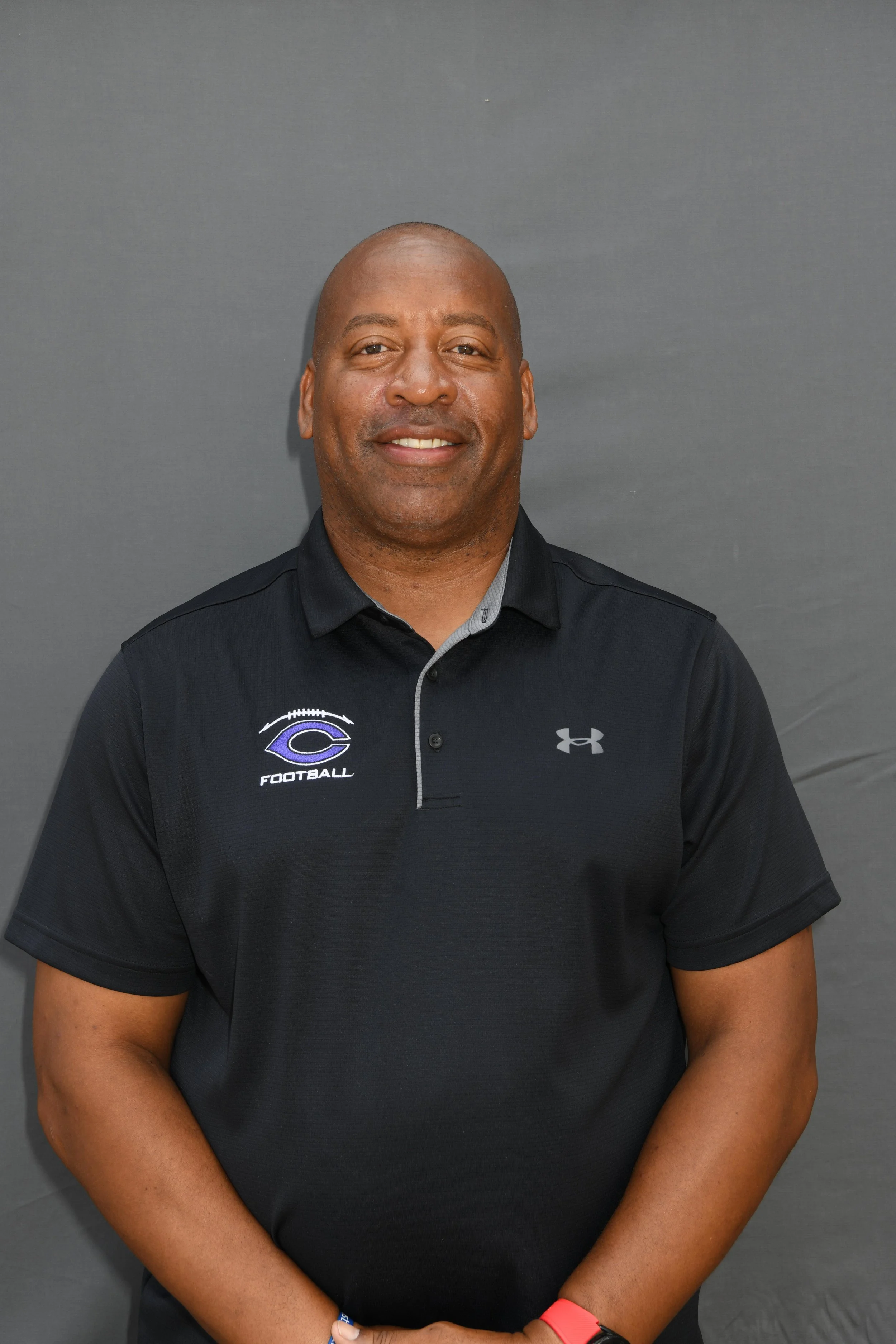 Man wearing a black polo shirt with a logo and the word 'football' on it, standing against a gray background, smiling, with arms crossed.